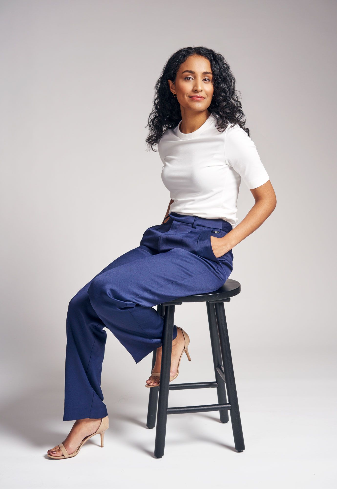Stylish woman in white top and navy wide-leg pants sitting on a black stool against a neutral background.
