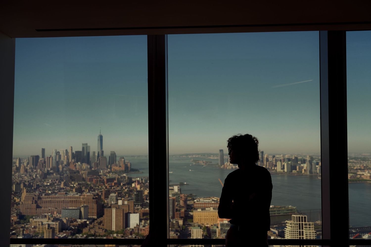 Silhouette of person gazing at New York City skyline with One World Trade Center visible through large windows.