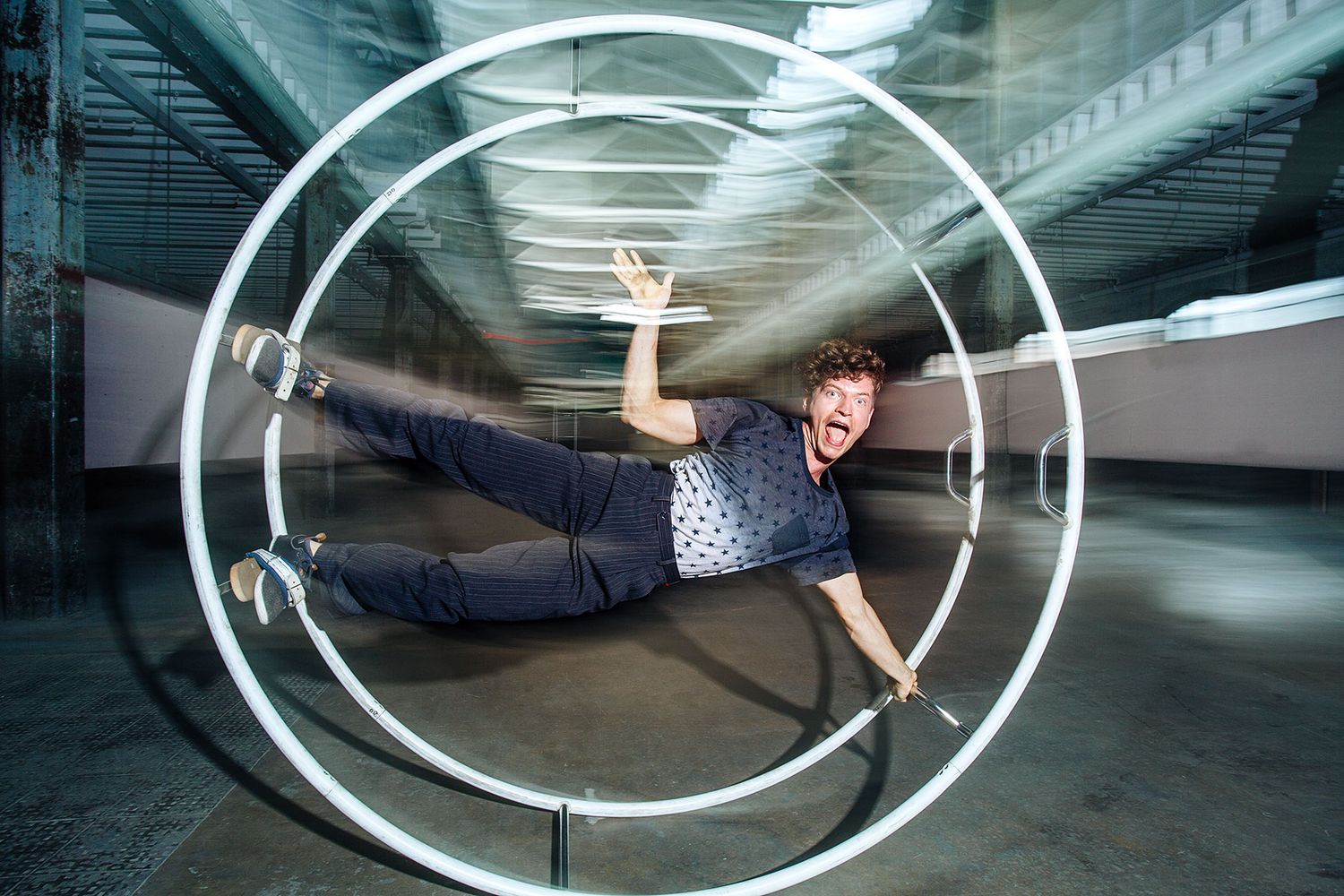 Person performing acrobatic stunt inside large circular metal rings in industrial warehouse space.