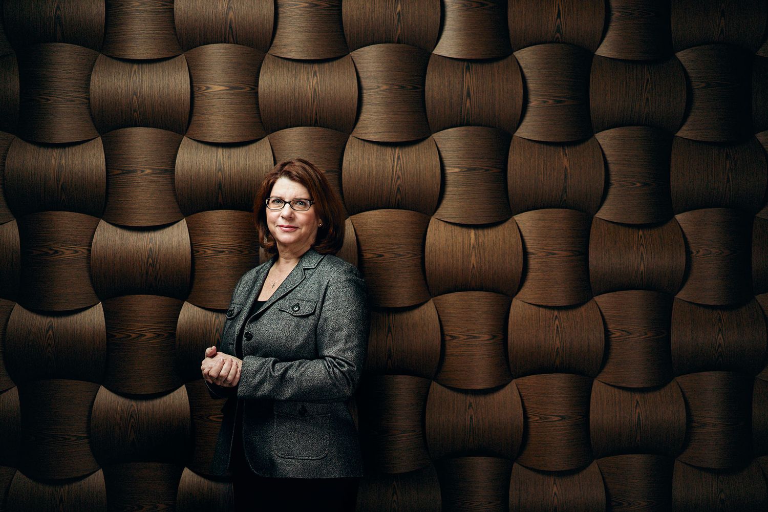Woman in gray blazer standing against textured wooden wall.