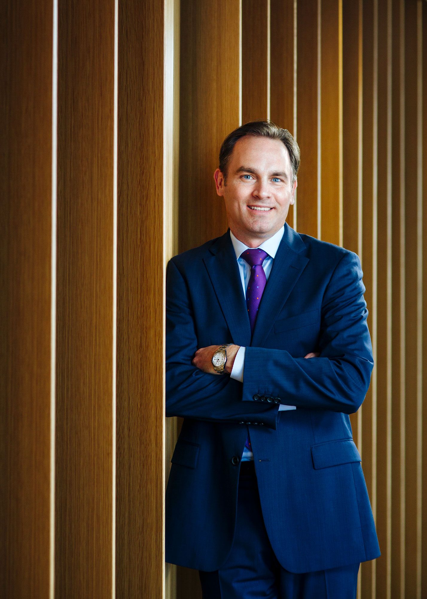 Confident businessman in blue suit and purple tie leans against wooden wall with arms crossed.