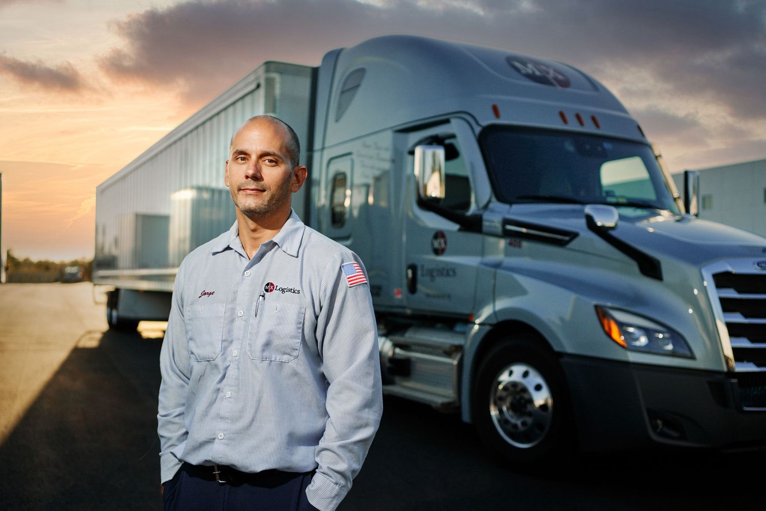 Male truck driver in uniform standing confidently in front of a semi-truck at sunset.