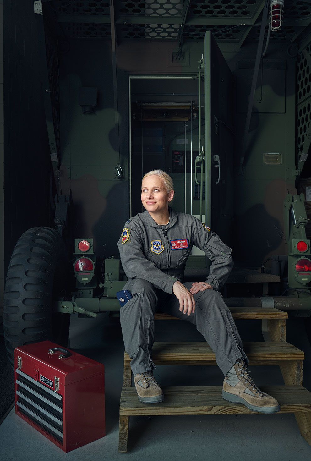 Female military aviator in flight suit sits on wooden steps beside a red toolbox near a military vehicle.