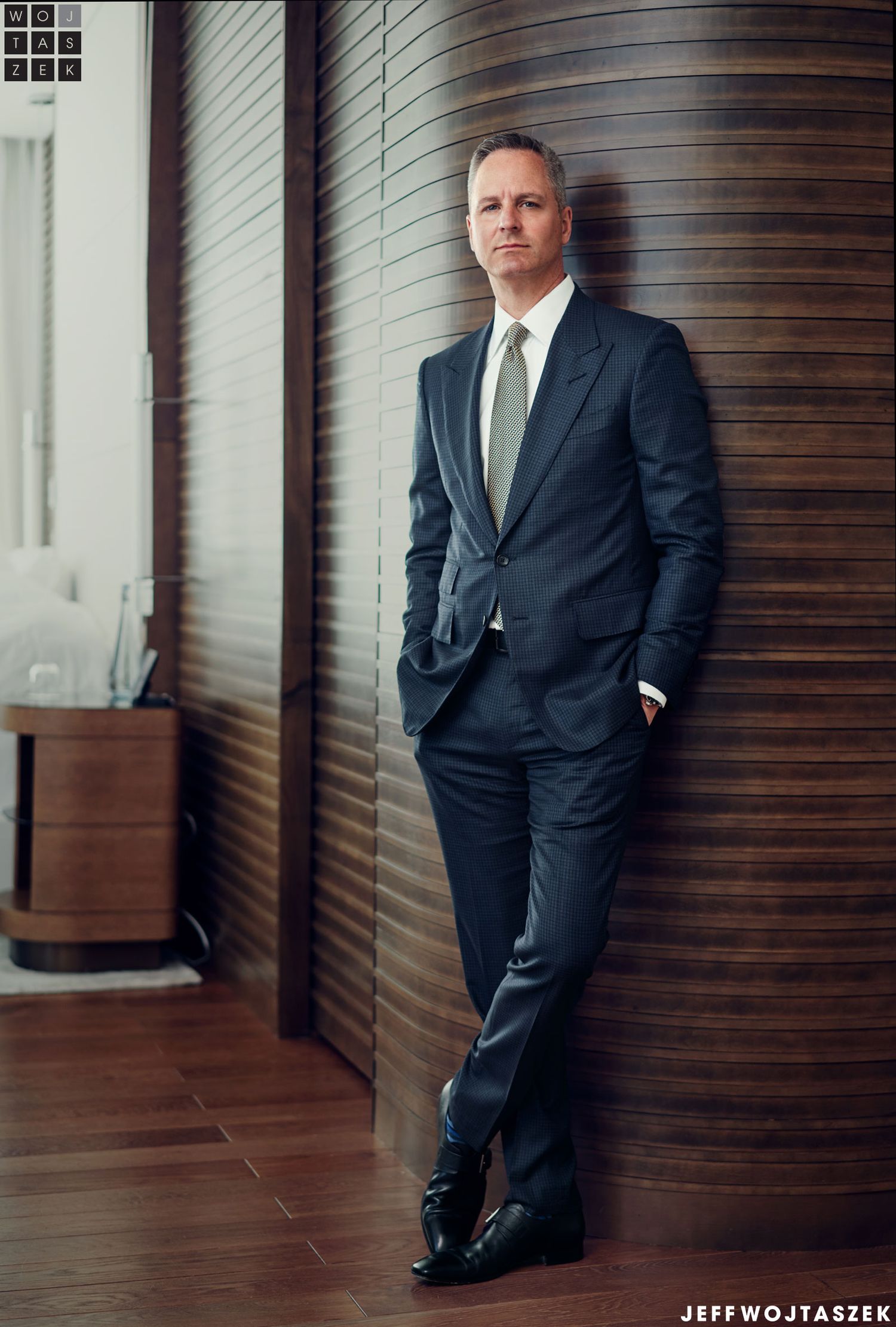 Professional man in navy pinstripe suit posing confidently in modern wood-paneled interior setting.