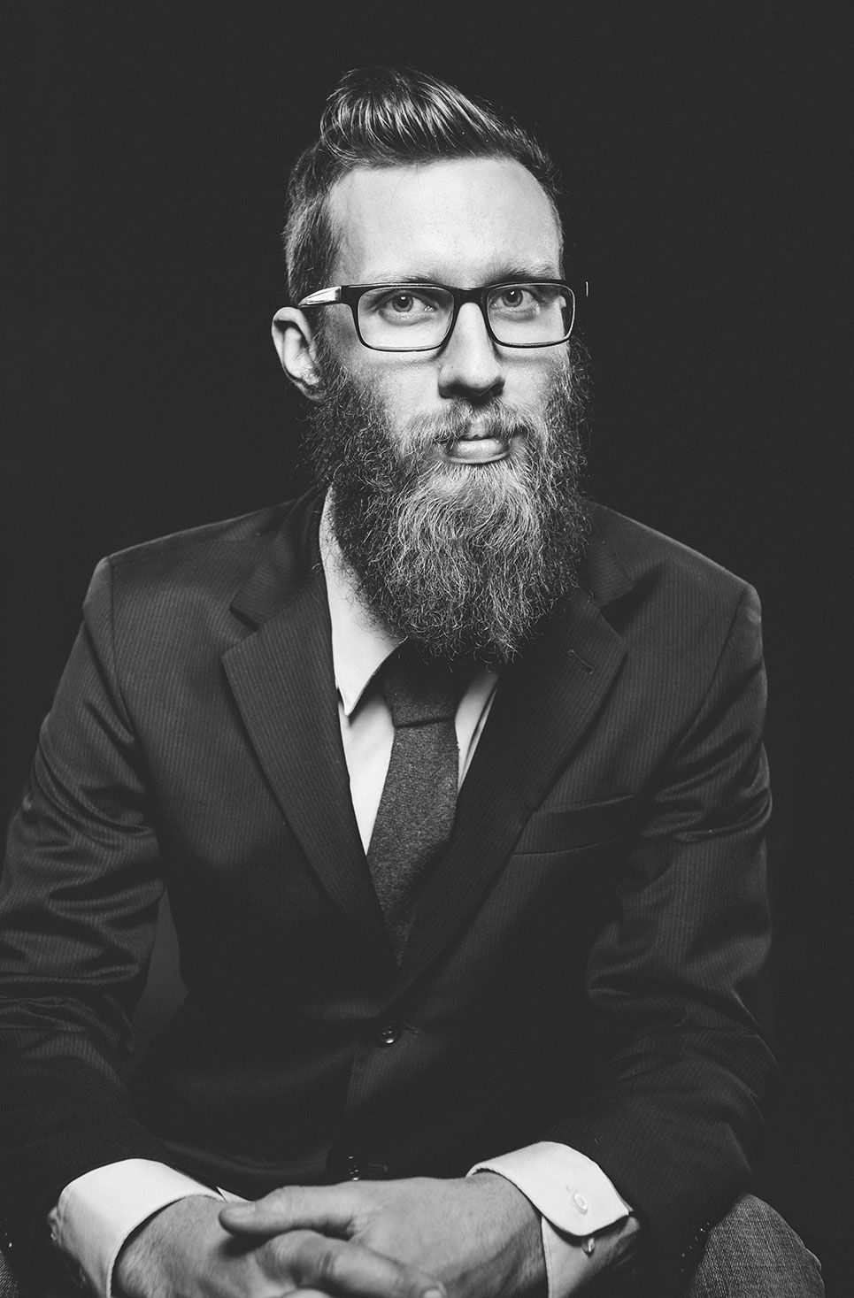 Black and white portrait of bearded man in suit and glasses with hands clasped.
