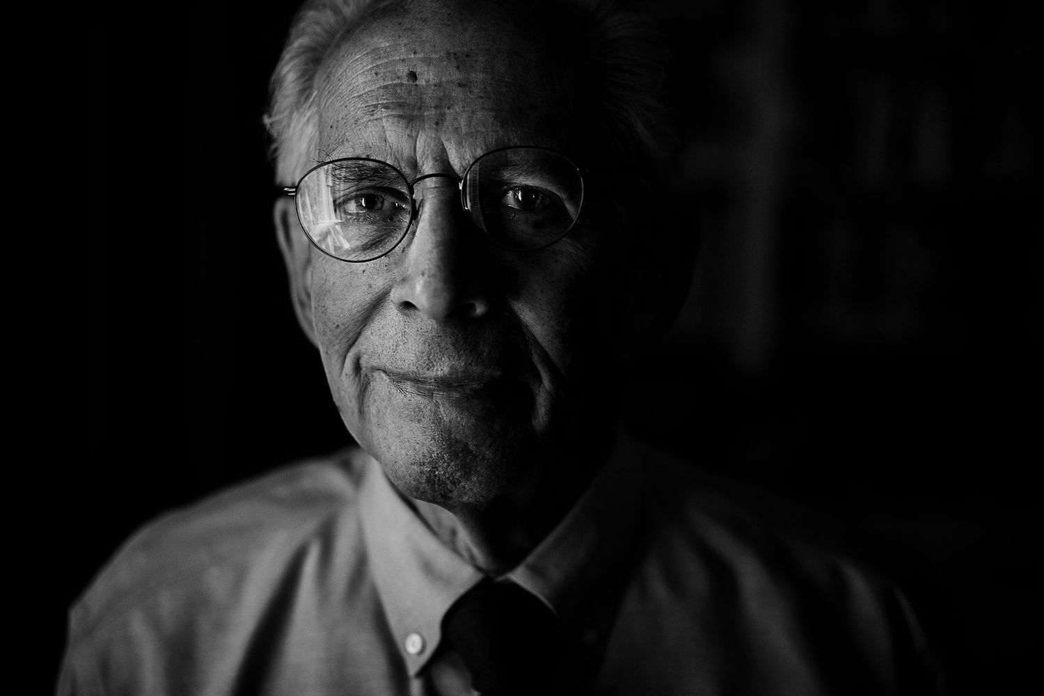 Black and white close-up portrait of elderly man wearing glasses in dim lighting.