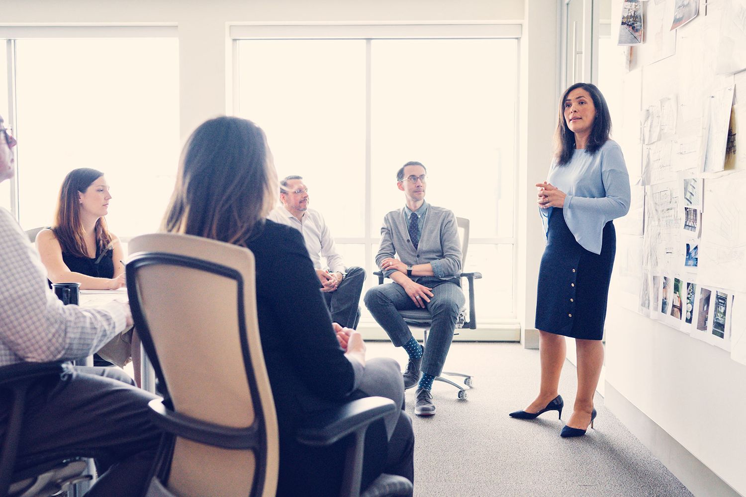 Female professional presents to seated colleagues in a modern office with design images on the wall.