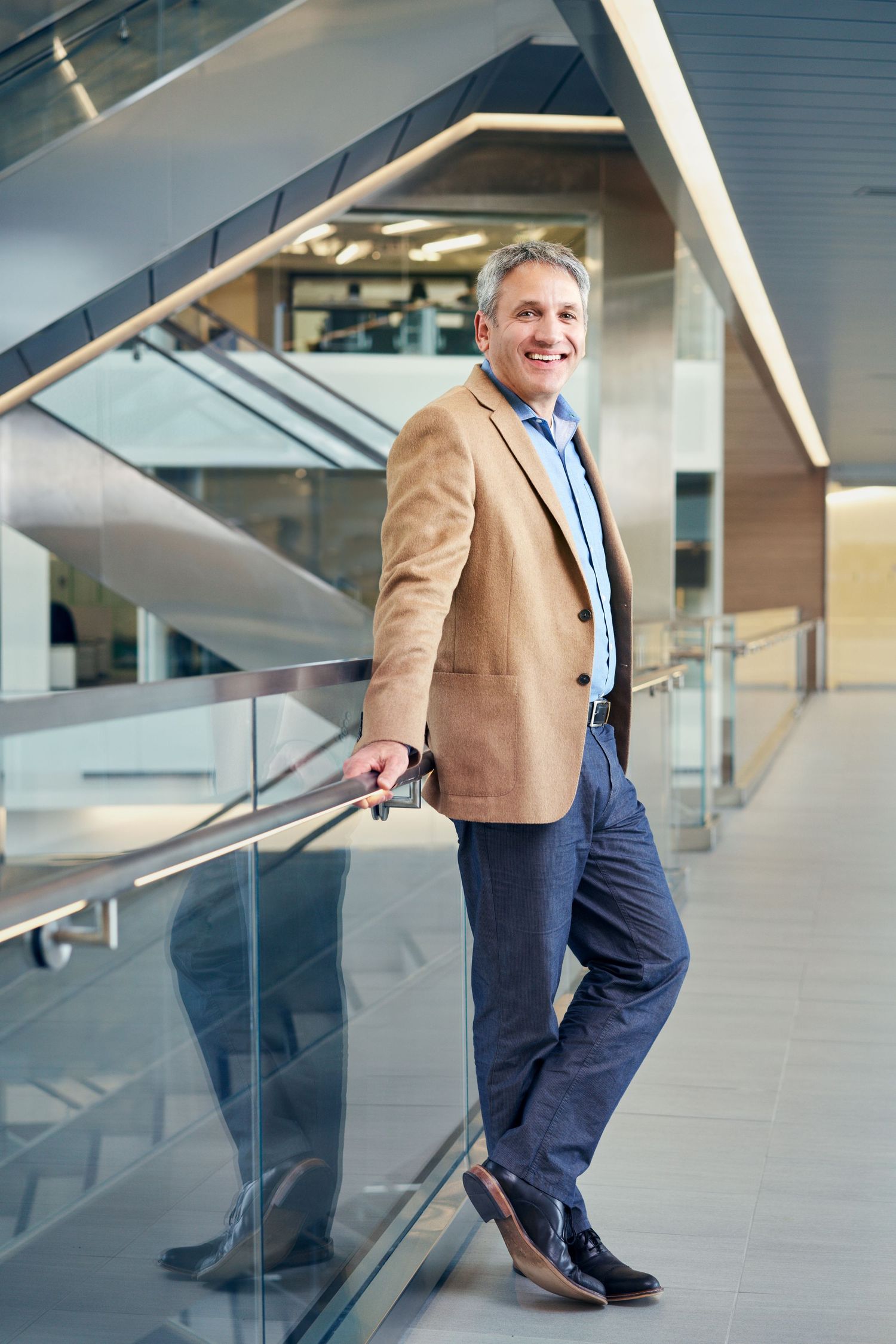 Smiling middle-aged businessman in beige blazer leaning on glass railing in modern office building.
