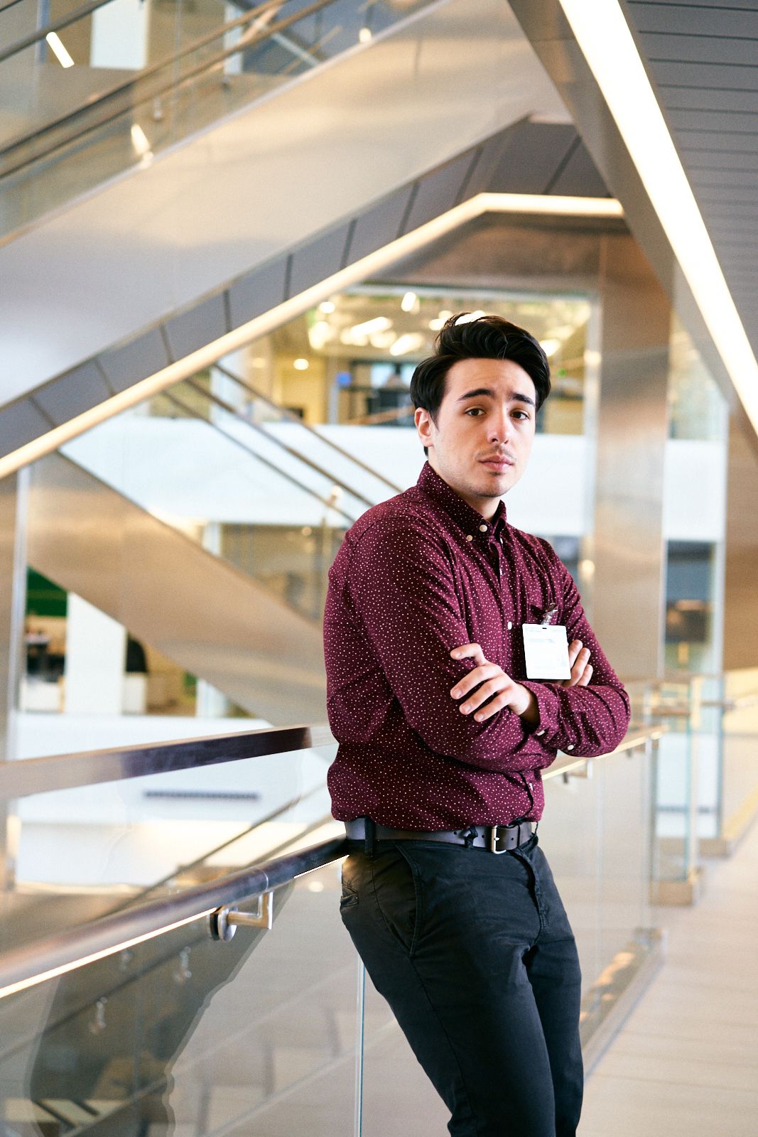 Young professional man in burgundy shirt with arms crossed, standing near glass railing in modern office building.