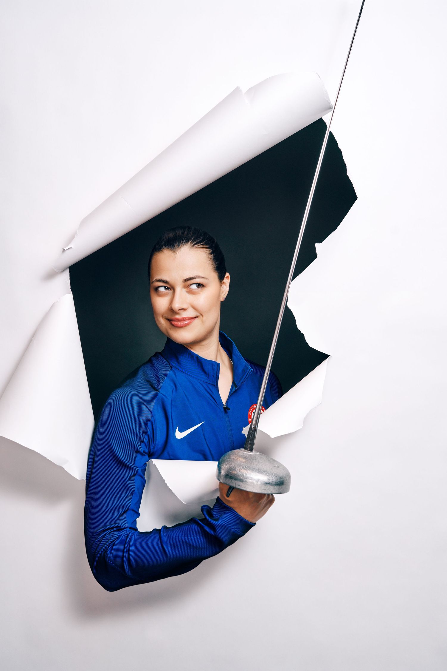 Female fencer in blue Nike jacket holds épée sword, emerging through torn white paper in creative sports portrait photo.