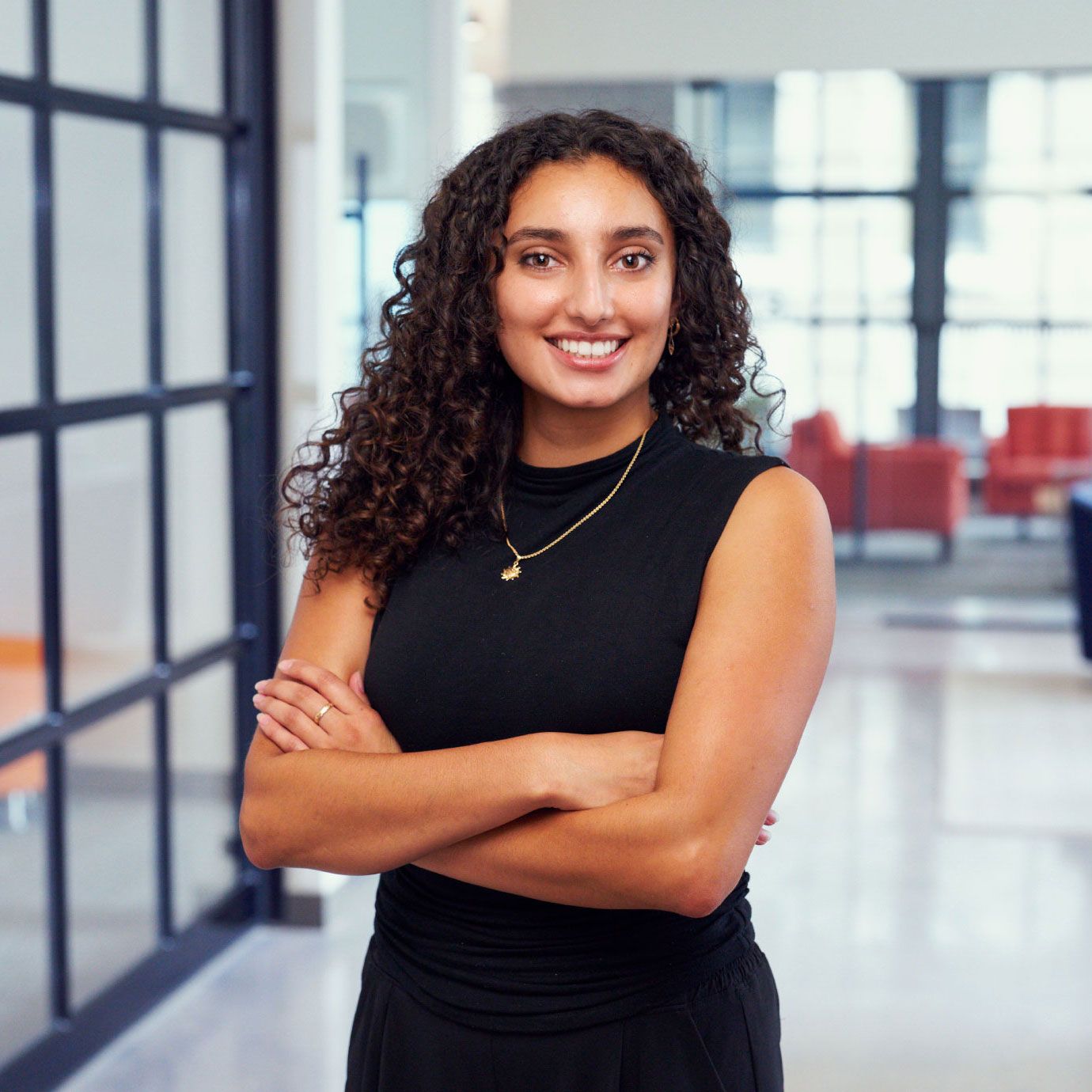 A young professional woman with curly hair smiles confidently with arms crossed in a modern office lobby.