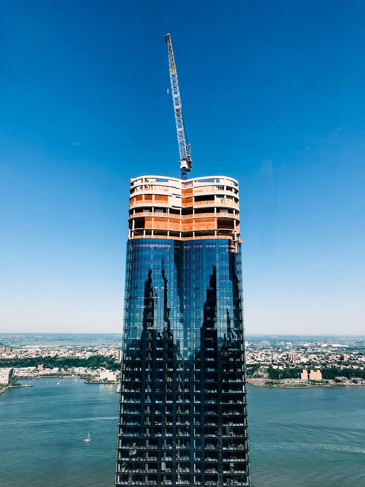 Aerial view of a skyscraper under construction with a crane on top, glass facade reflecting city, waterfront visible below.