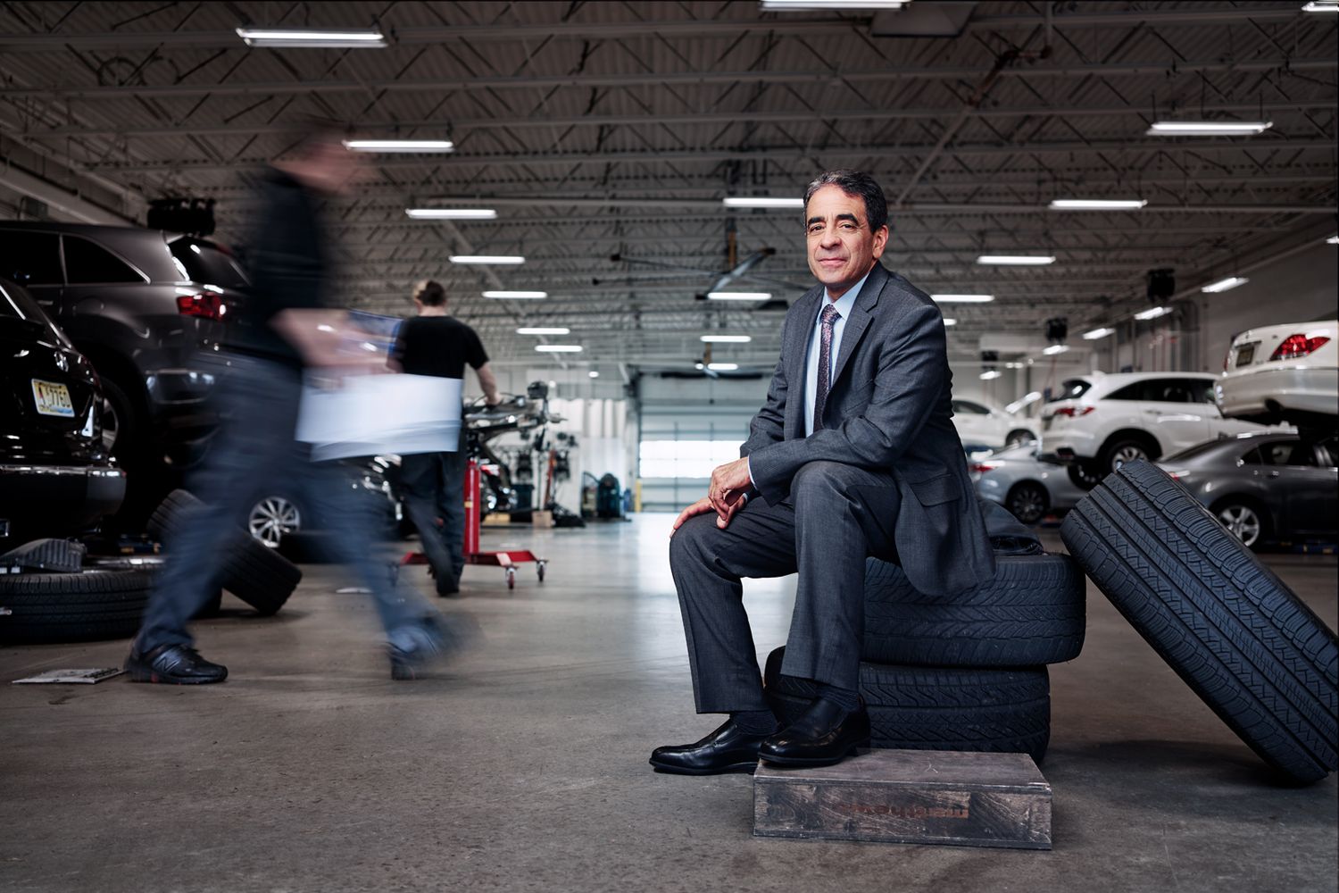 Businessman in suit sitting on tires in an auto repair shop with mechanics working in background.