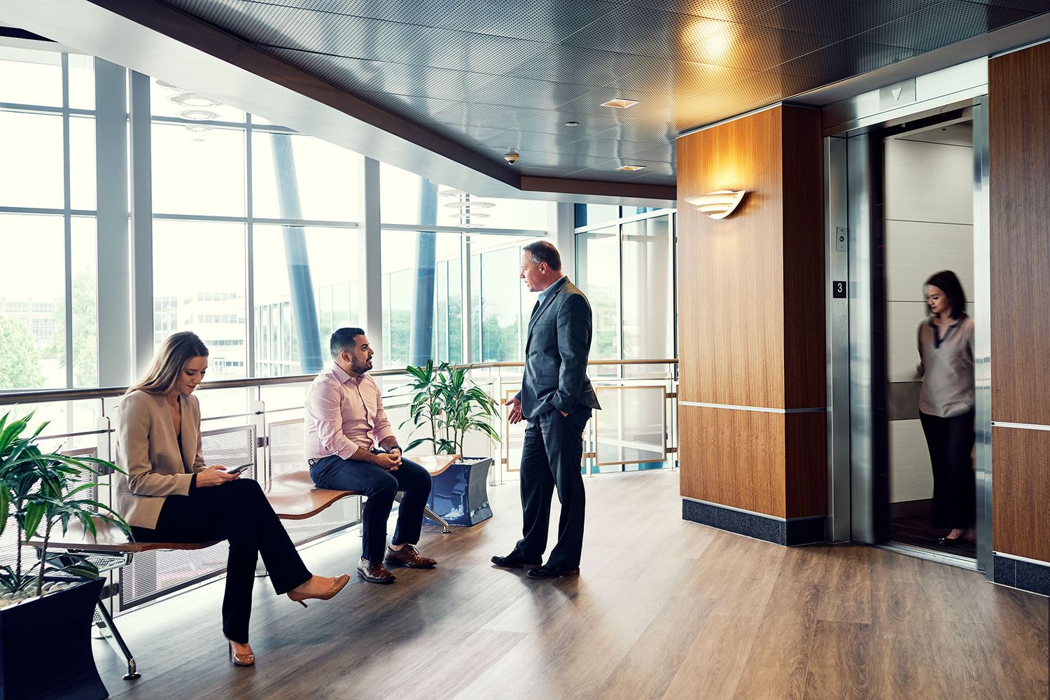 Professionals networking in a modern office lobby with wood floors, large windows, and an elevator.