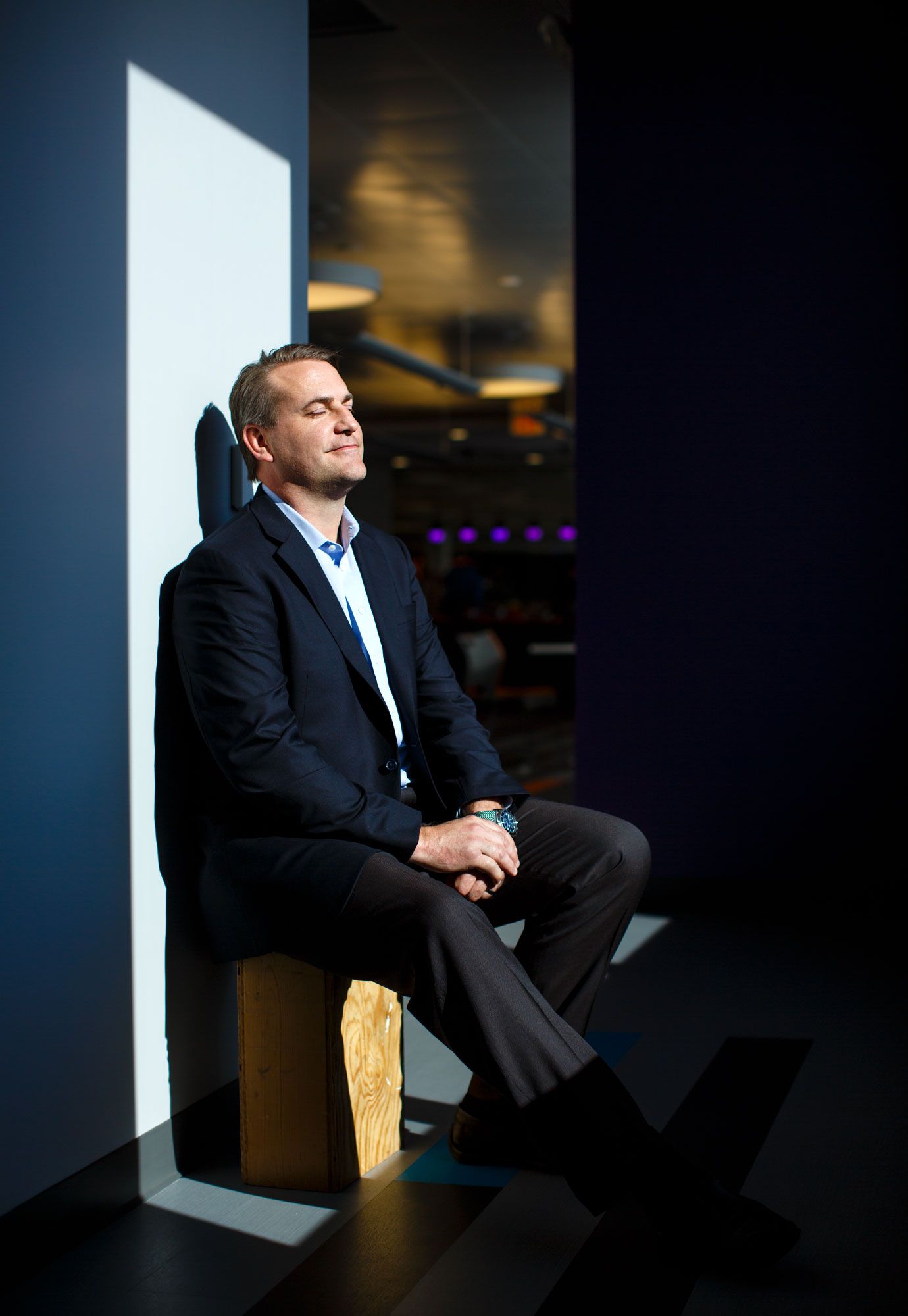 Man in navy blazer sits relaxed in dramatic light, eyes closed, in a modern blue-lit interior space.