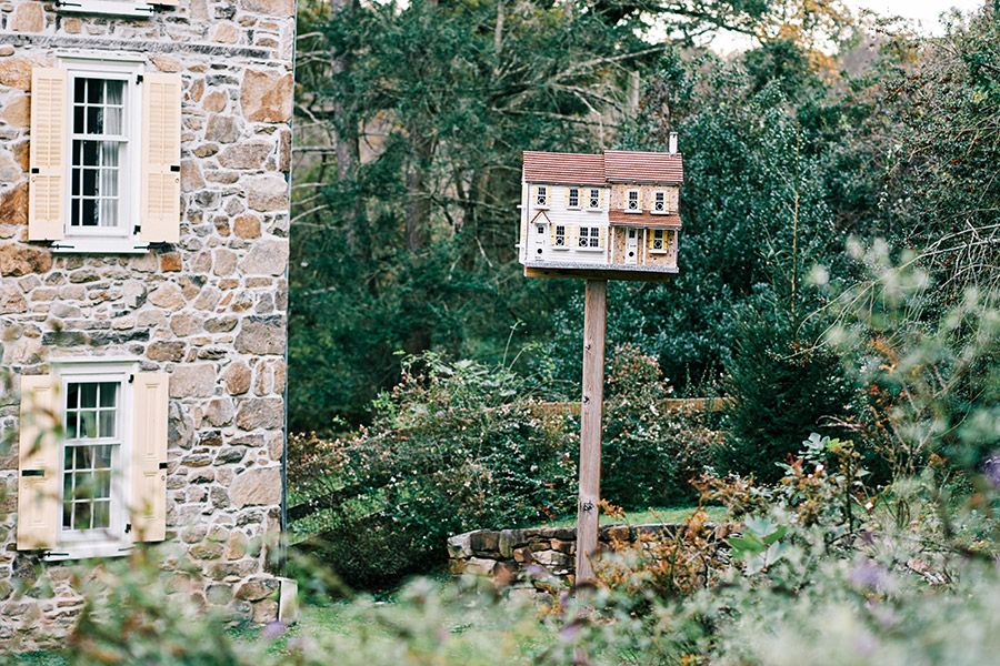 A decorative wooden birdhouse shaped like a colonial home on a post surrounded by lush green gardens near a stone building.