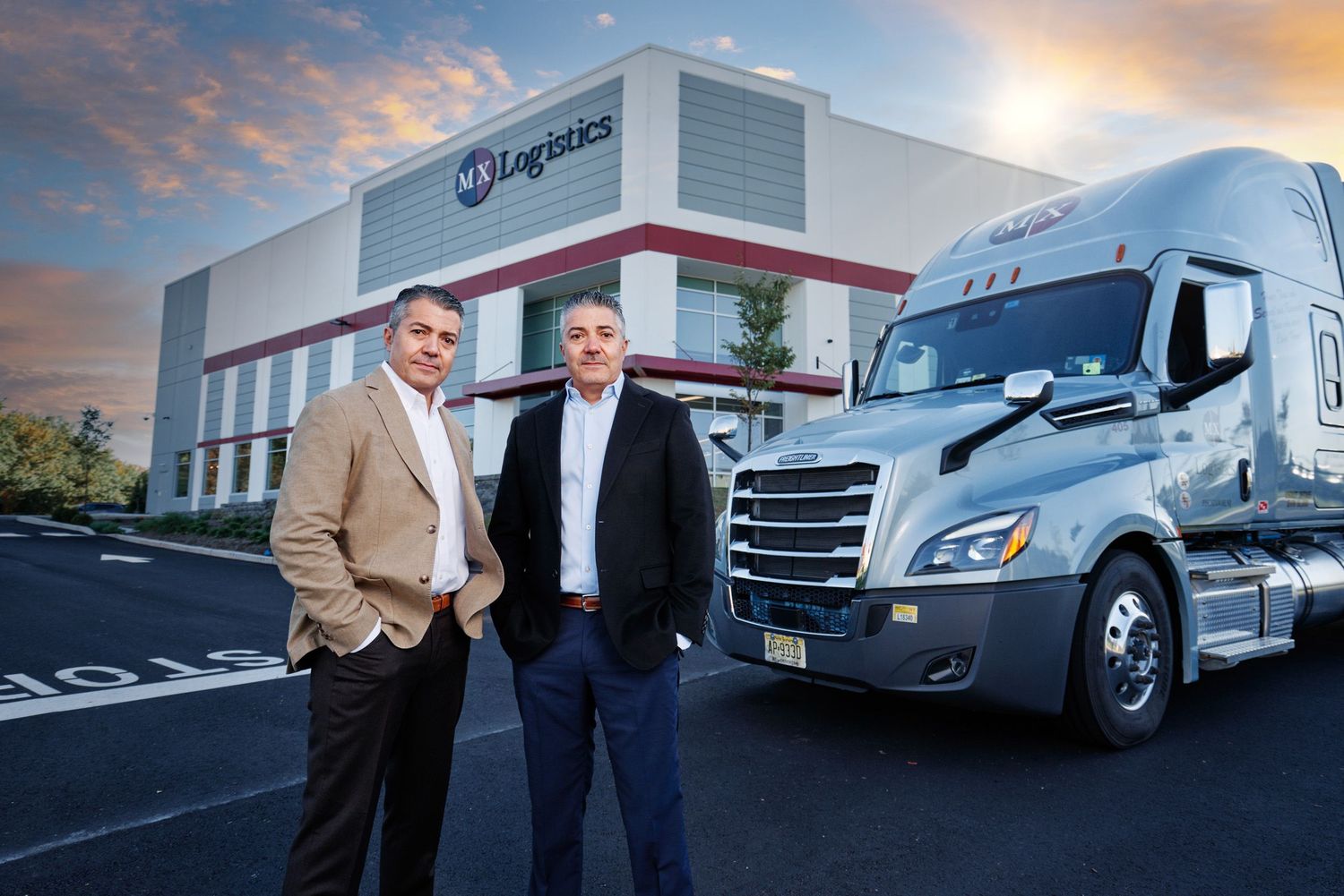 Two logistics company executives posing in front of semi-trucks at a distribution facility.