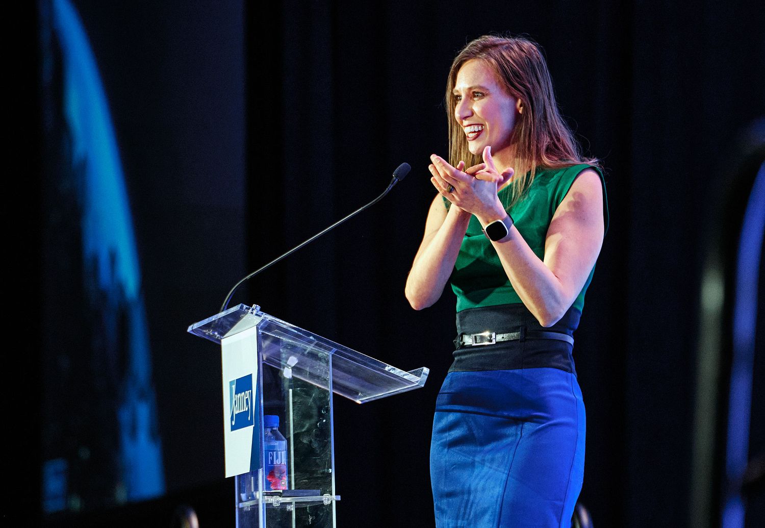 Woman in green top and blue skirt speaking at a glass podium during a professional event.