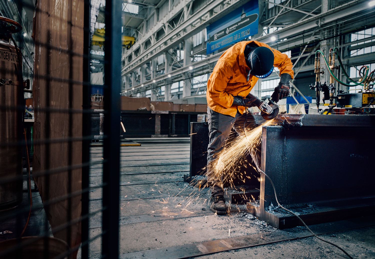 Worker in orange protective gear grinding metal with bright sparks flying in industrial facility.
