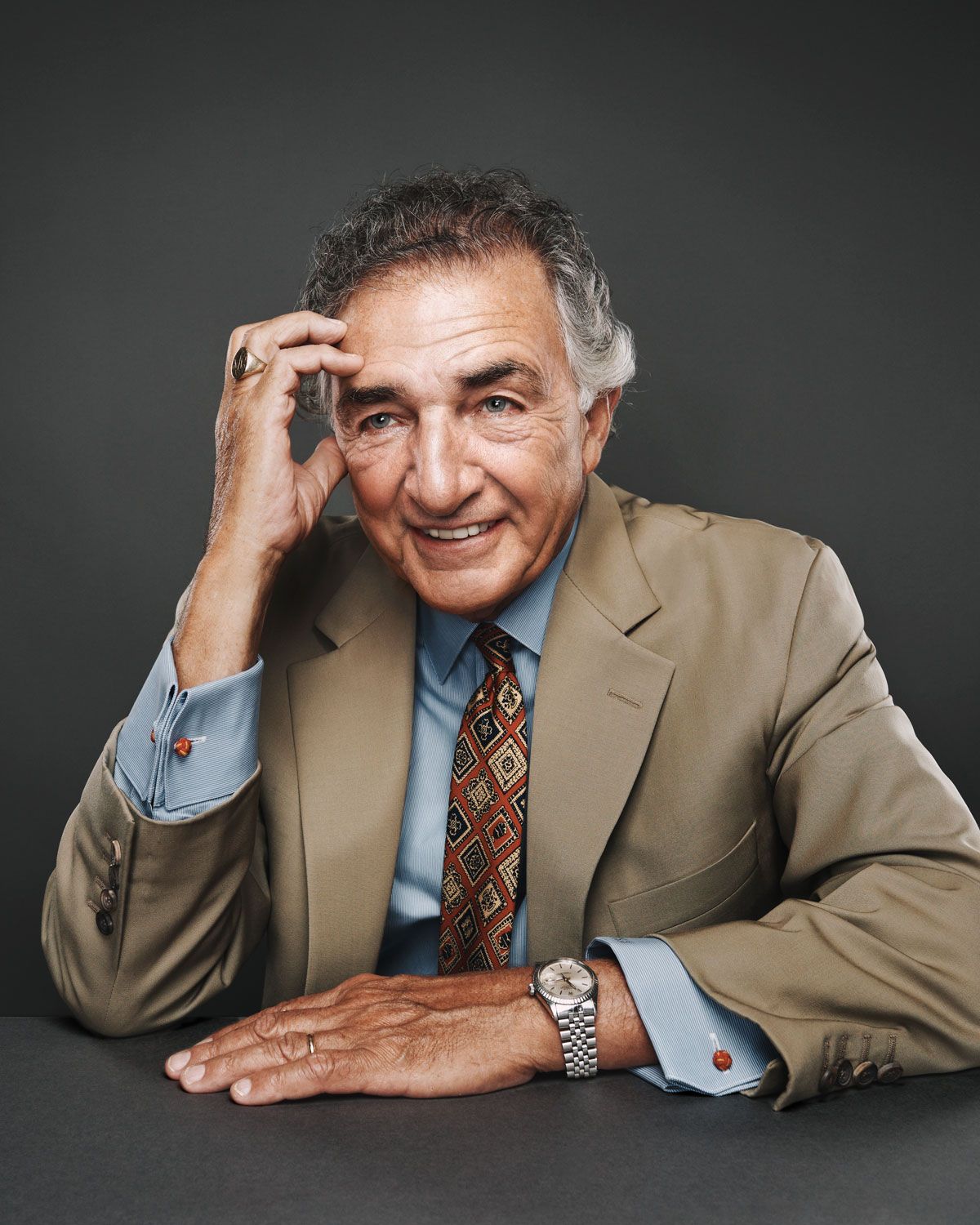 Smiling elderly man in beige suit and patterned tie poses confidently at a table against gray background.