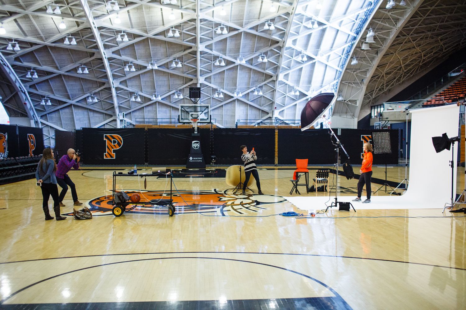 A photography crew sets up lighting equipment on a basketball court with orange and black team logos during an indoor photo shoot.