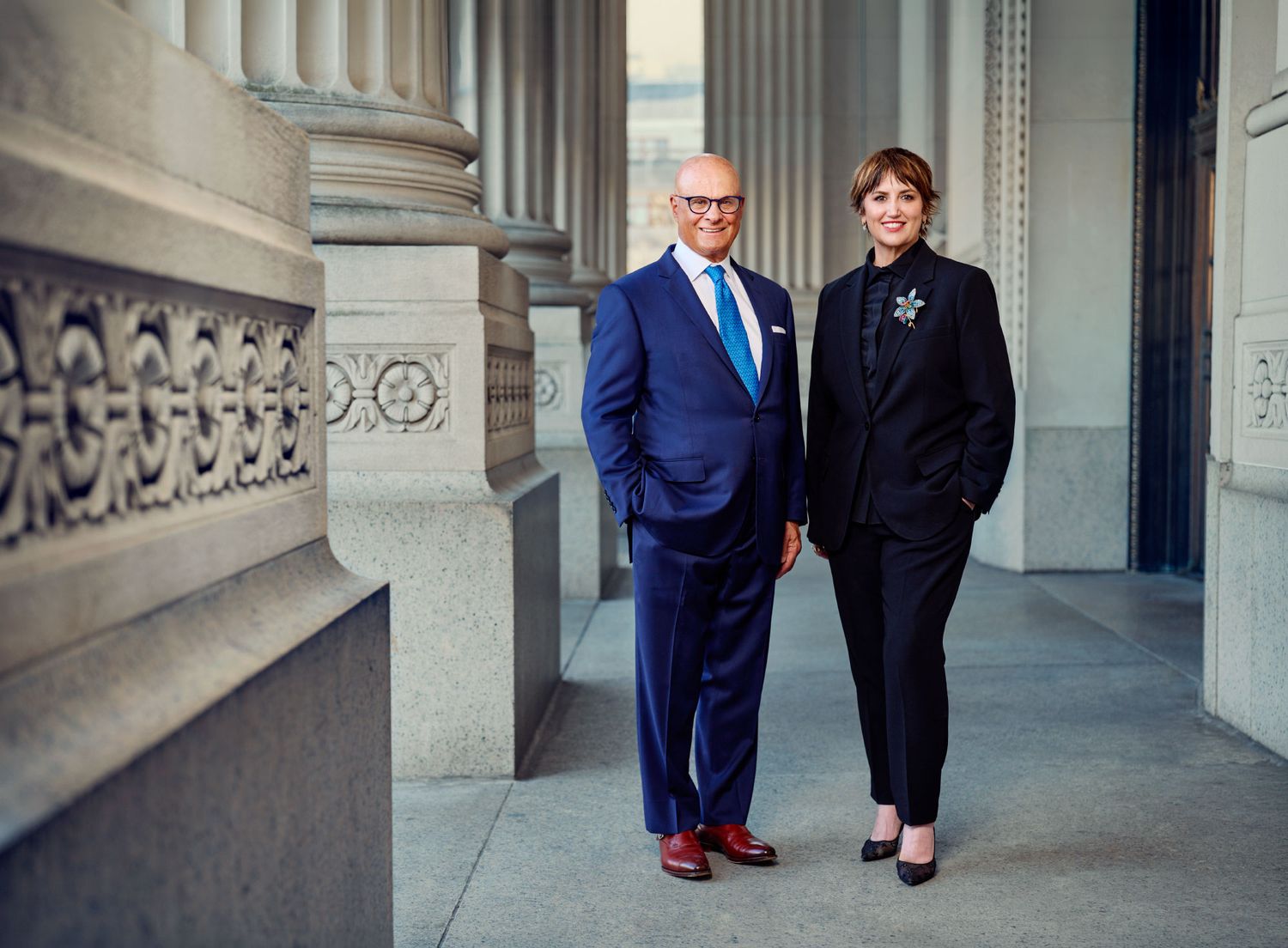 Two business professionals in formal attire pose together beside large stone columns.