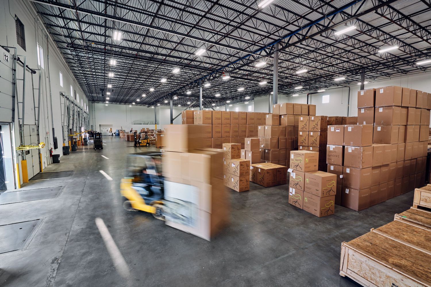 Forklift moving cardboard boxes in a large warehouse with high ceilings and stacked pallets.