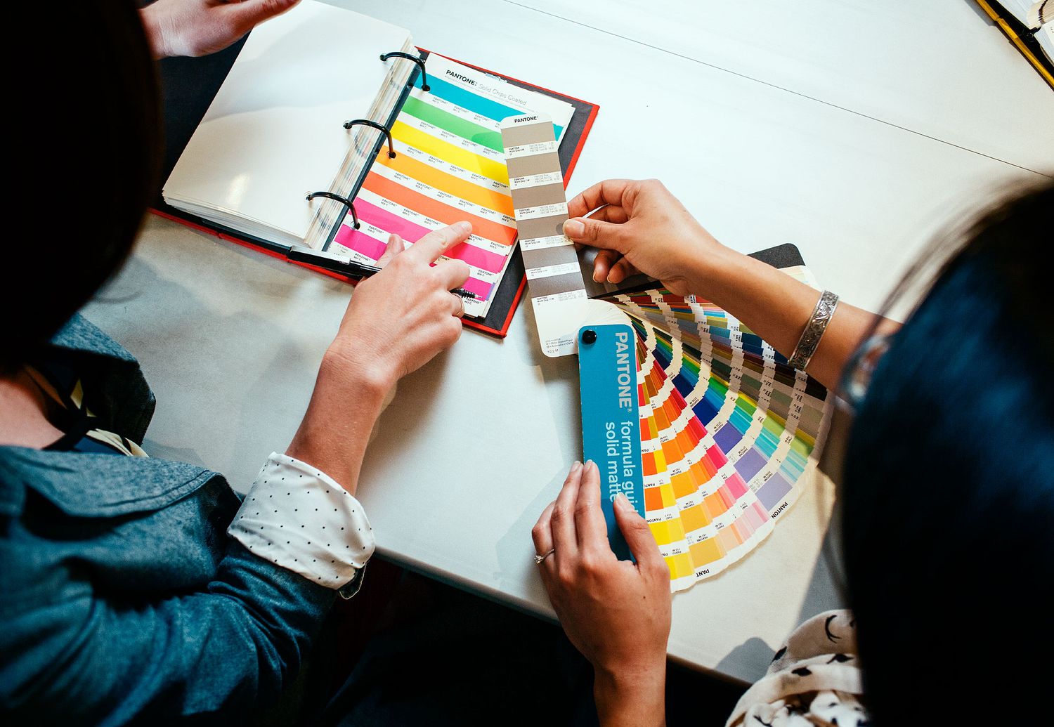 Designers compare colorful Pantone color swatches at a desk during a creative design meeting.