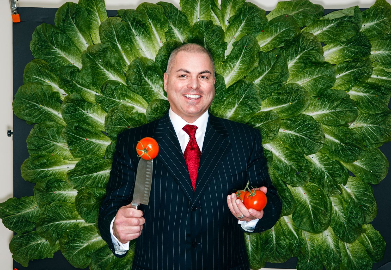 Cheerful man in black suit holds tomatoes in front of large decorative lettuce wall display.
