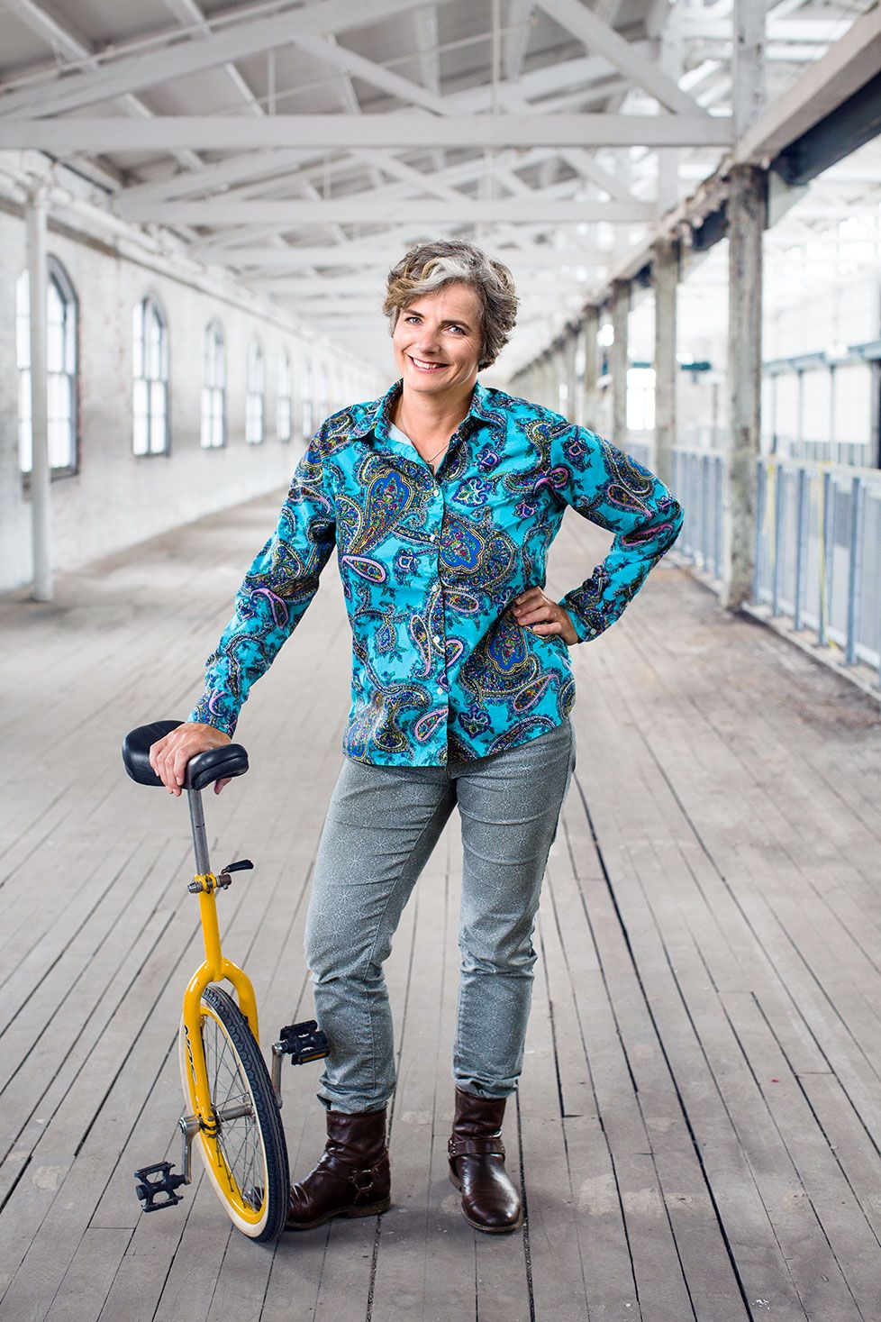 Woman in colorful paisley shirt posing with a unicycle indoors.