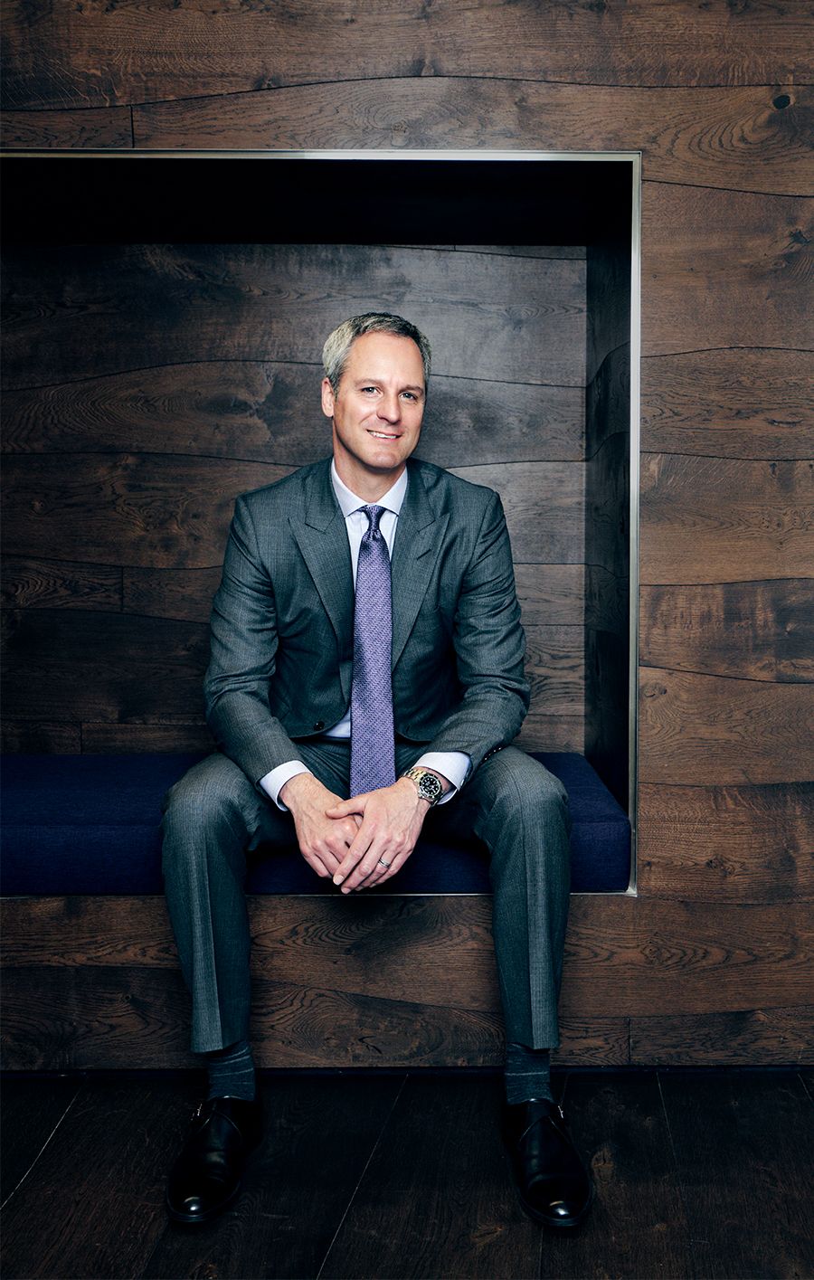 Professional man in gray suit and purple tie sitting confidently on a blue bench against a wood panel wall.