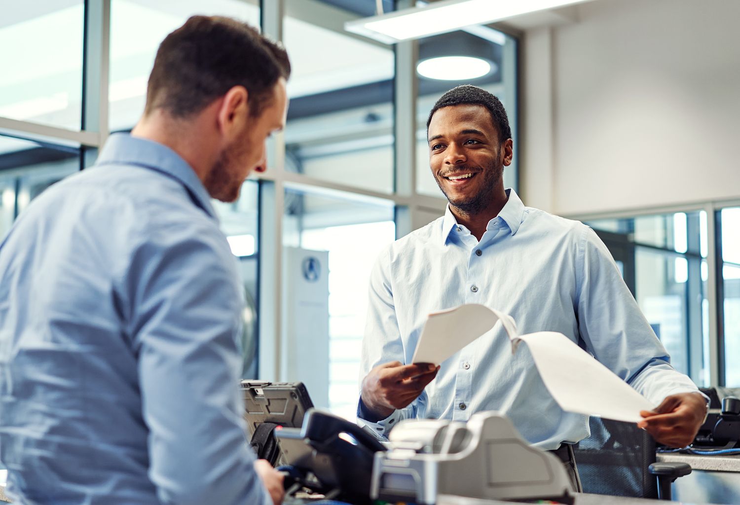 Smiling businessman assisting a customer at a modern office reception desk with paperwork and equipment.