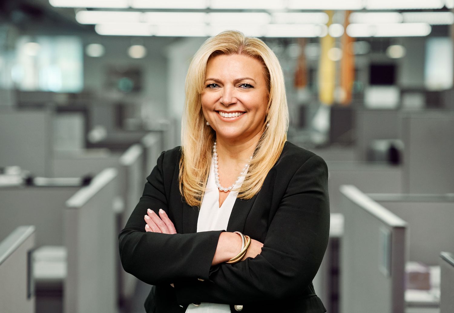 Smiling blonde businesswoman in black blazer with arms crossed in modern office.