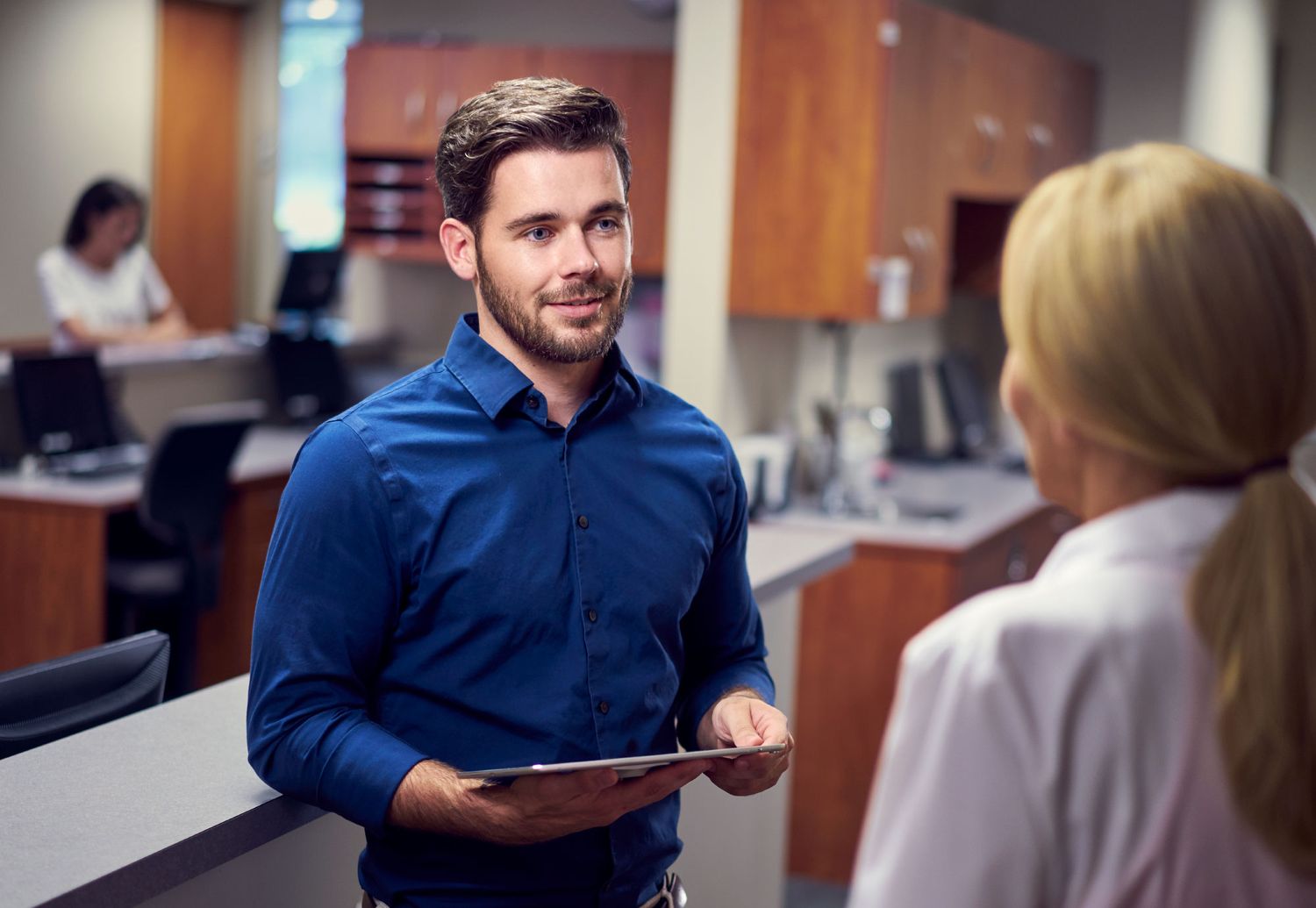 Young man in blue shirt holding clipboard speaking with blonde woman at medical reception desk.
