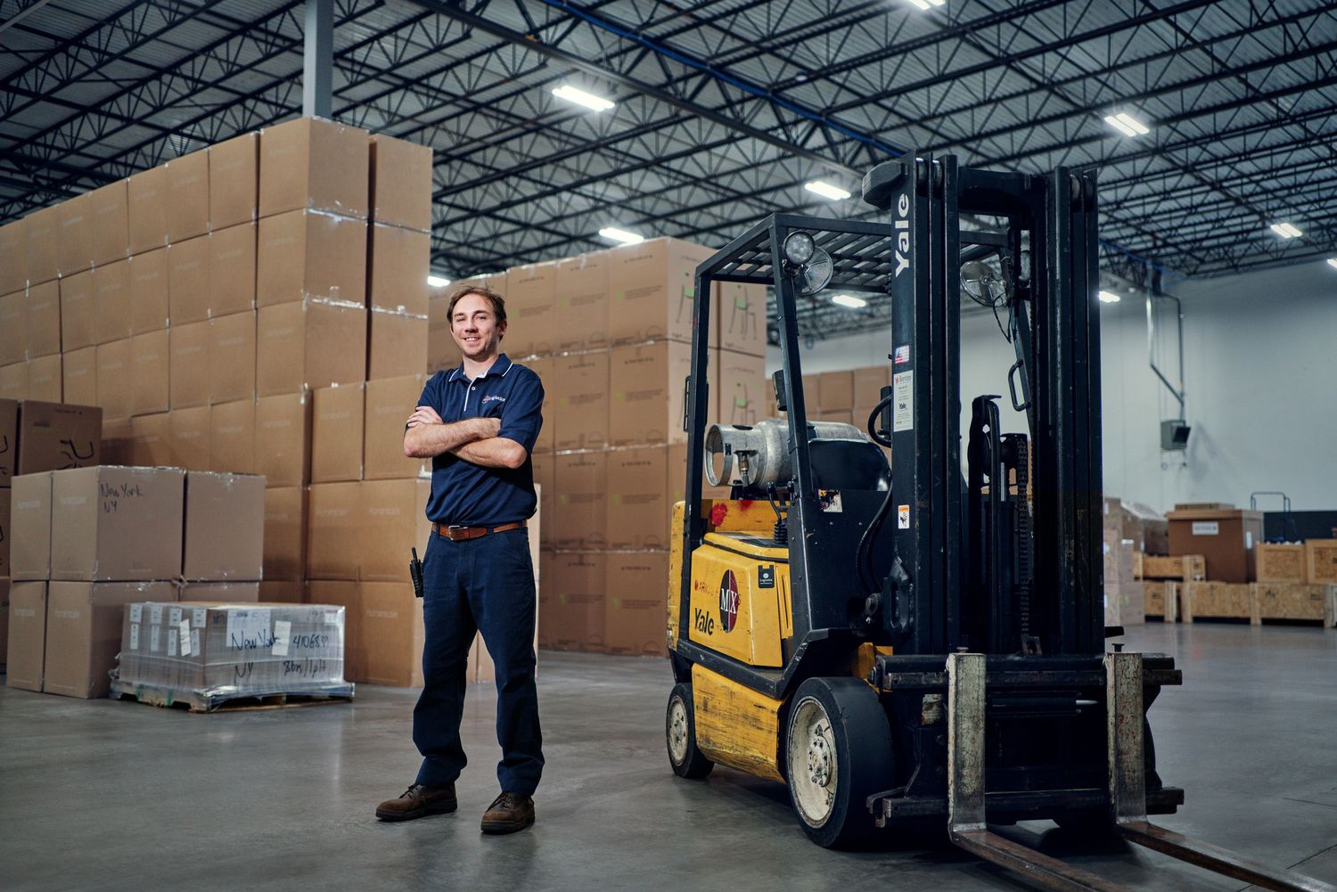 Smiling warehouse worker standing beside a yellow forklift in a large distribution center.