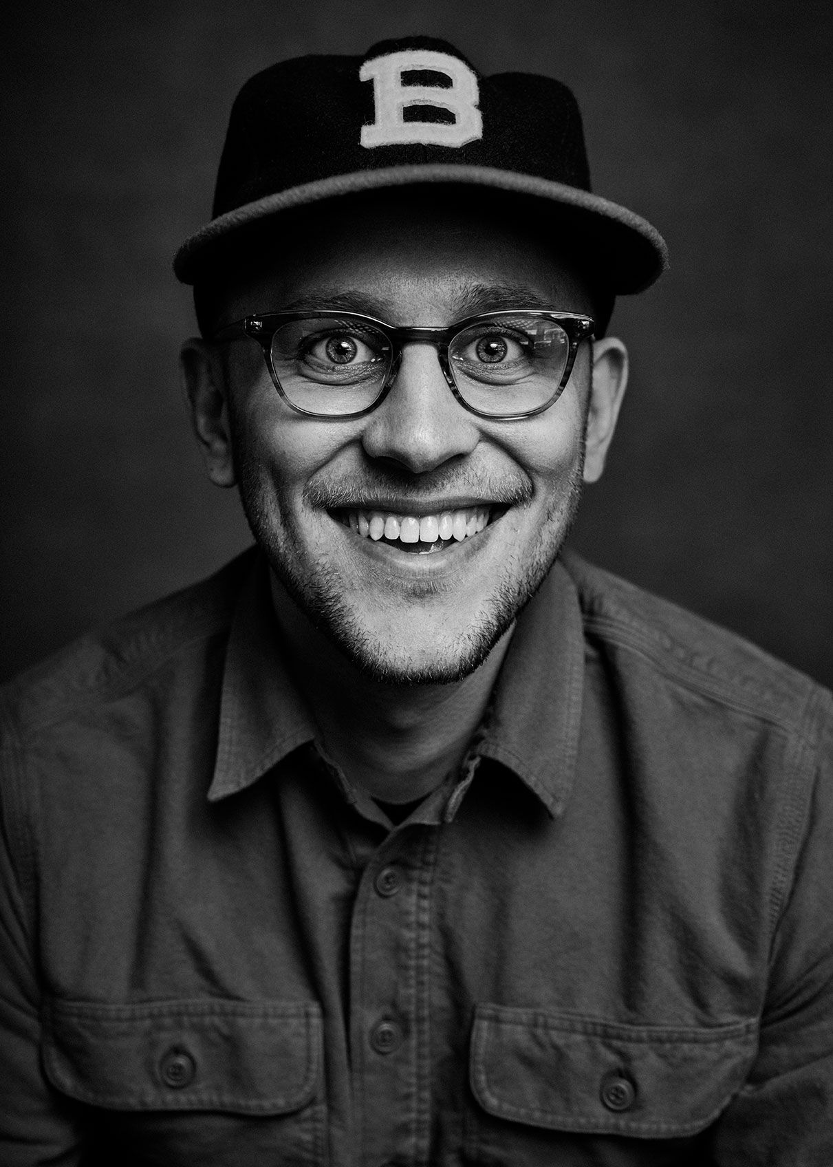 Smiling man wearing glasses and a B baseball cap in a black and white portrait photo.