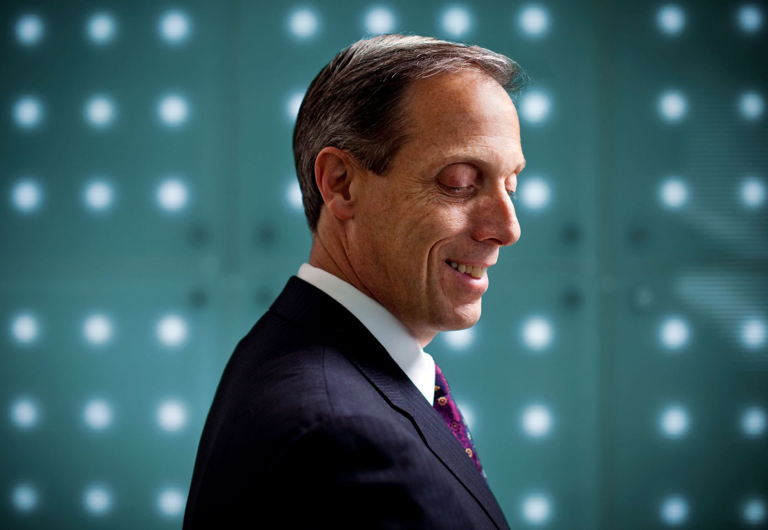 Professional businessman in dark suit smiles confidently against illuminated green background.