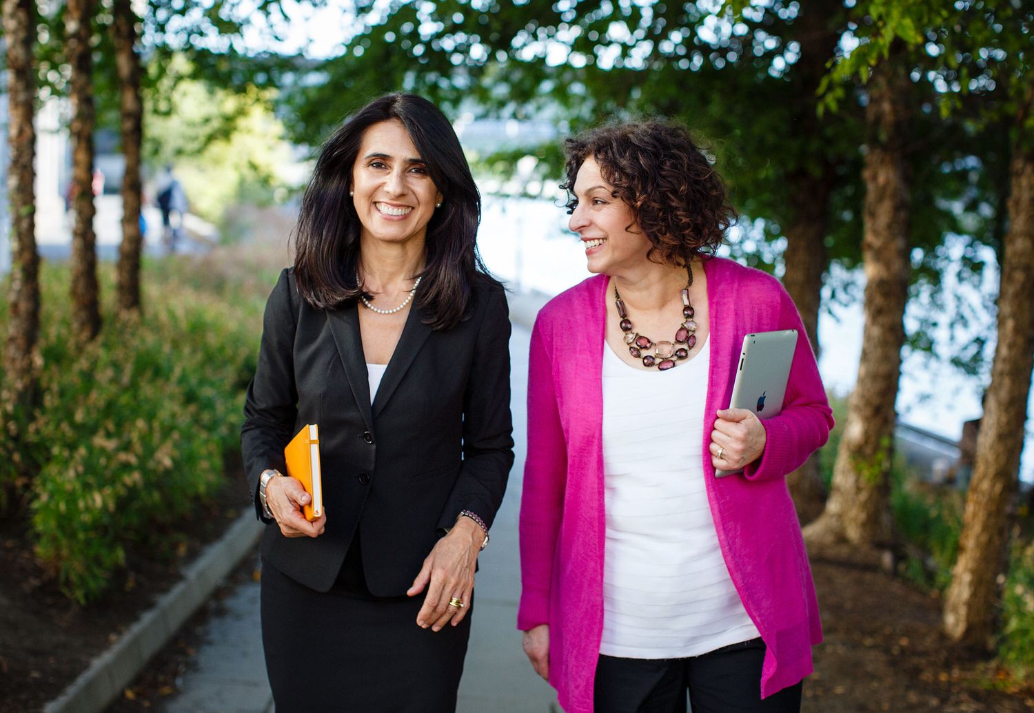 Two professional women walk and talk together along a tree-lined outdoor pathway.