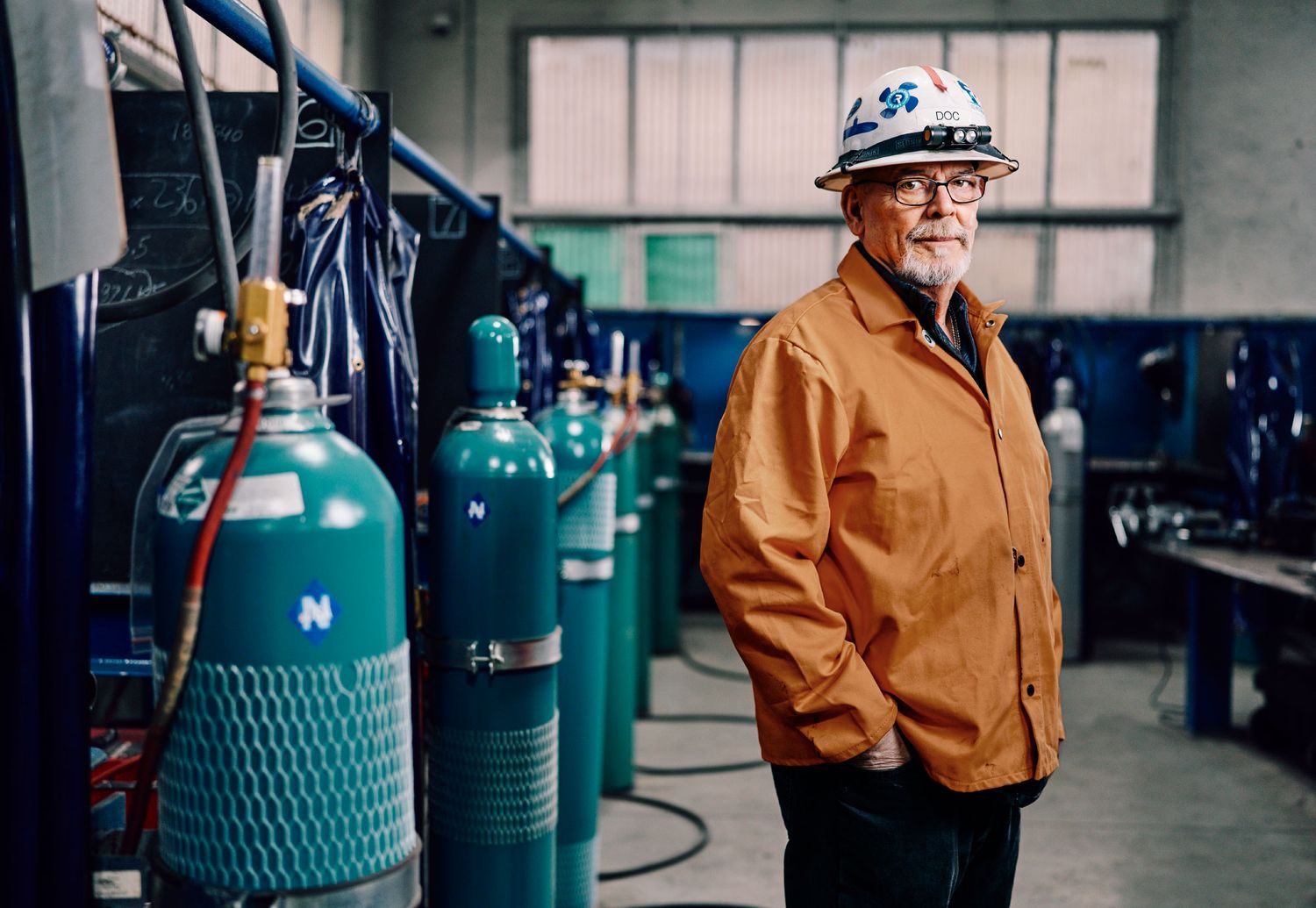 Worker in orange jacket and hard hat stands beside industrial gas cylinders in a manufacturing facility.