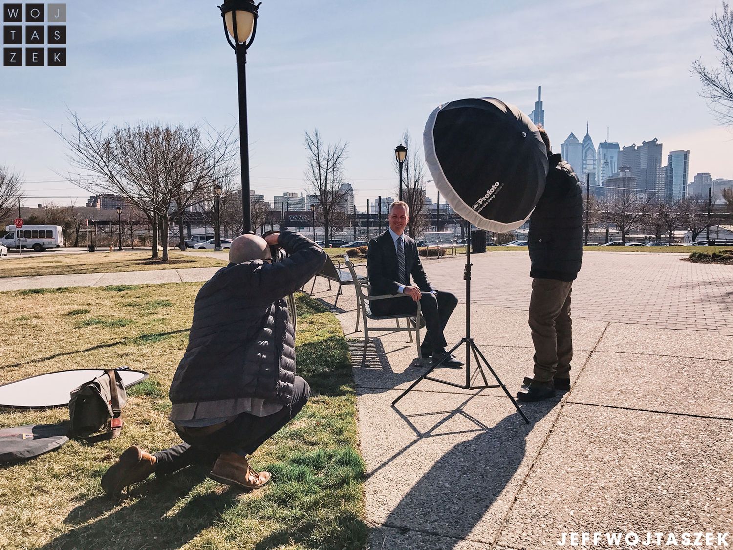 Outdoor professional photo shoot with city skyline backdrop, photographer, subject in chair, and large reflector on sunny day.