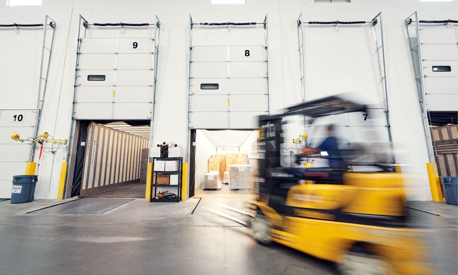Yellow forklift in motion inside a busy warehouse loading dock with numbered bay doors.