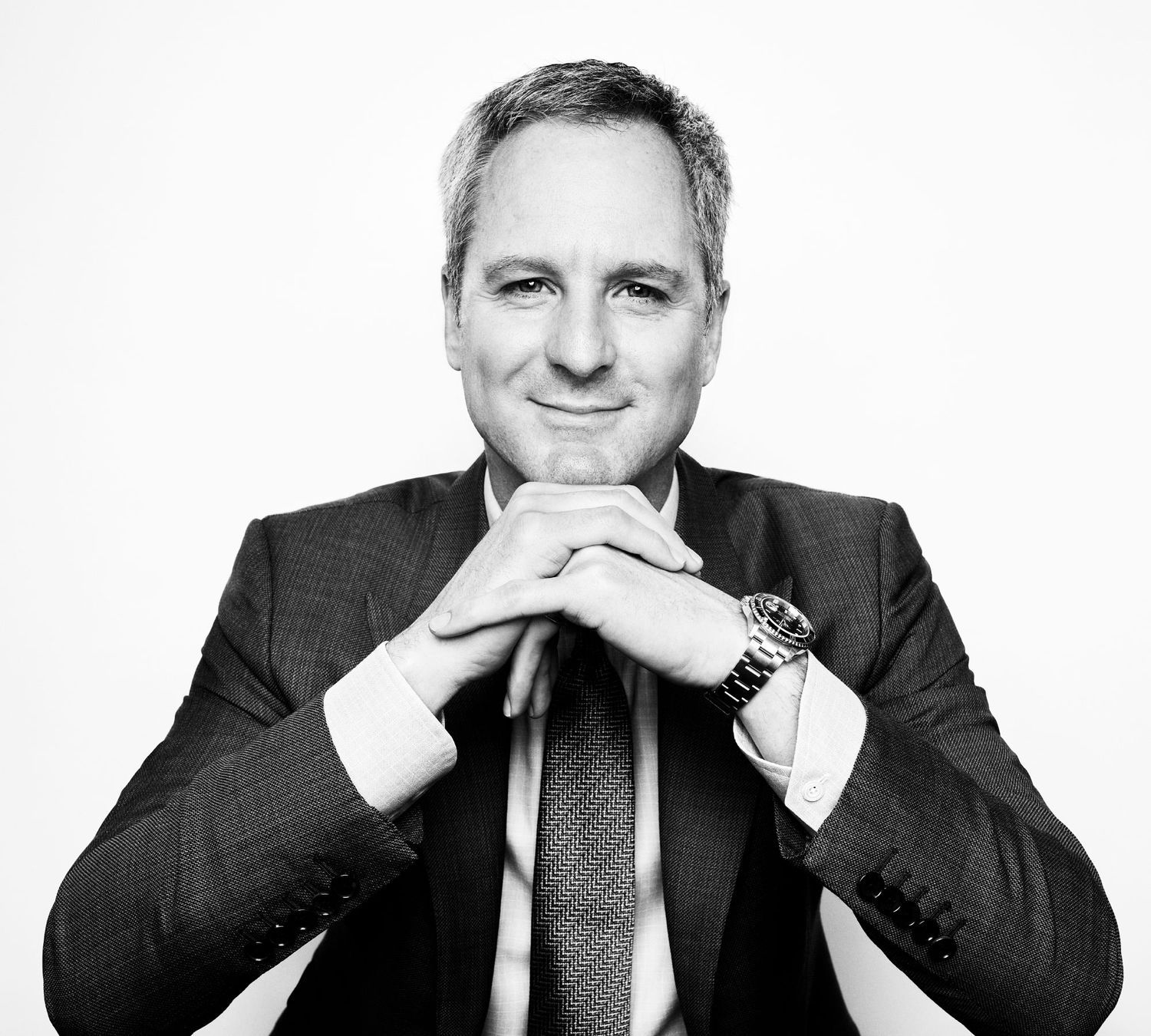 Smiling executive in dark suit with hands clasped in black and white professional portrait.