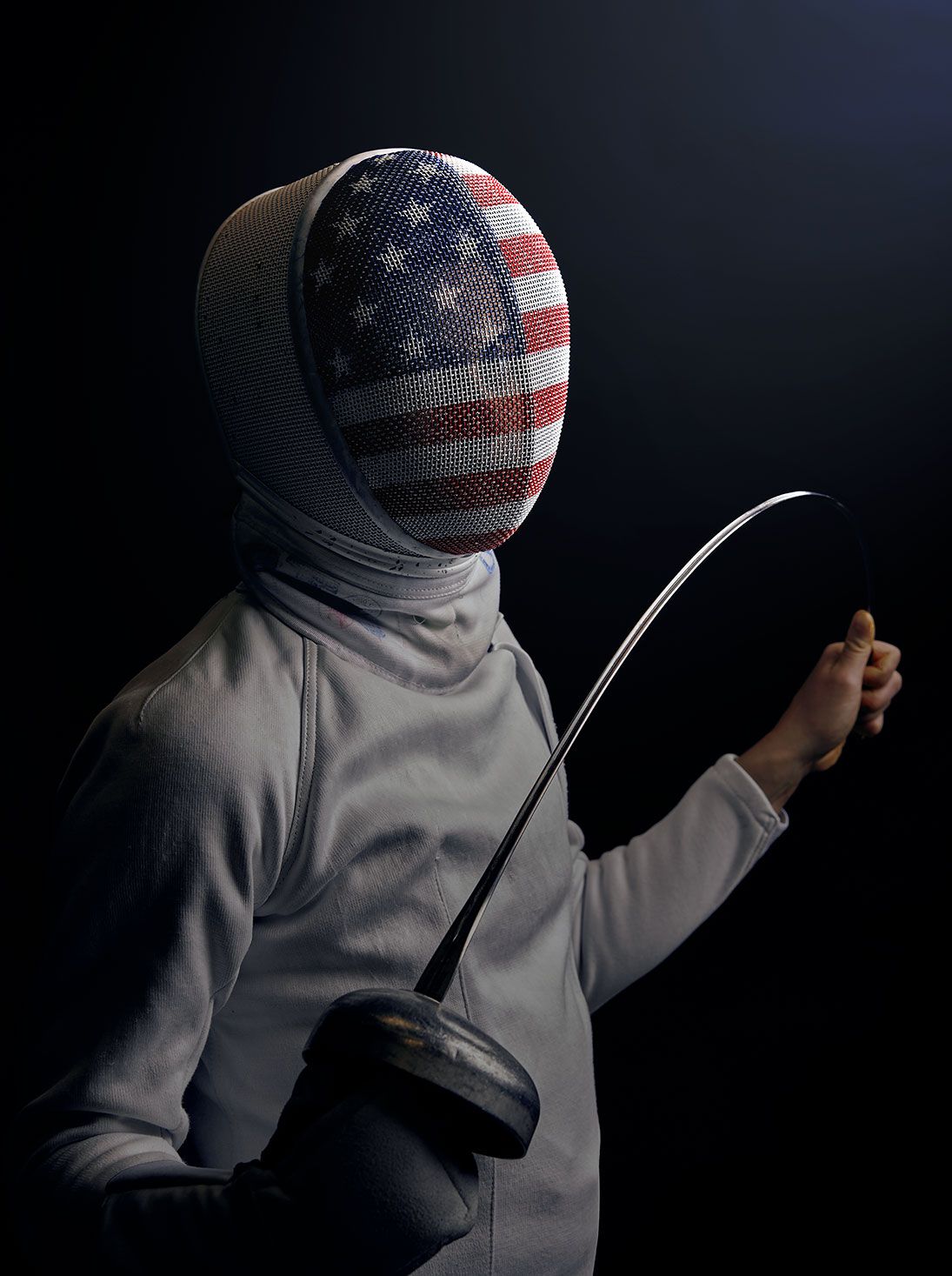 American fencer in white uniform holding épée with USA flag helmet on dark background.