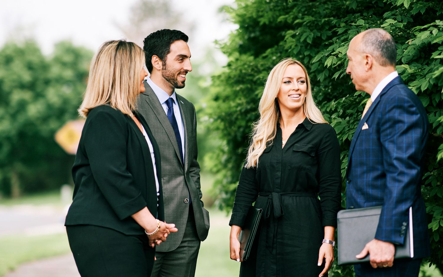 Four business professionals networking outdoors in formal attire near lush green trees.