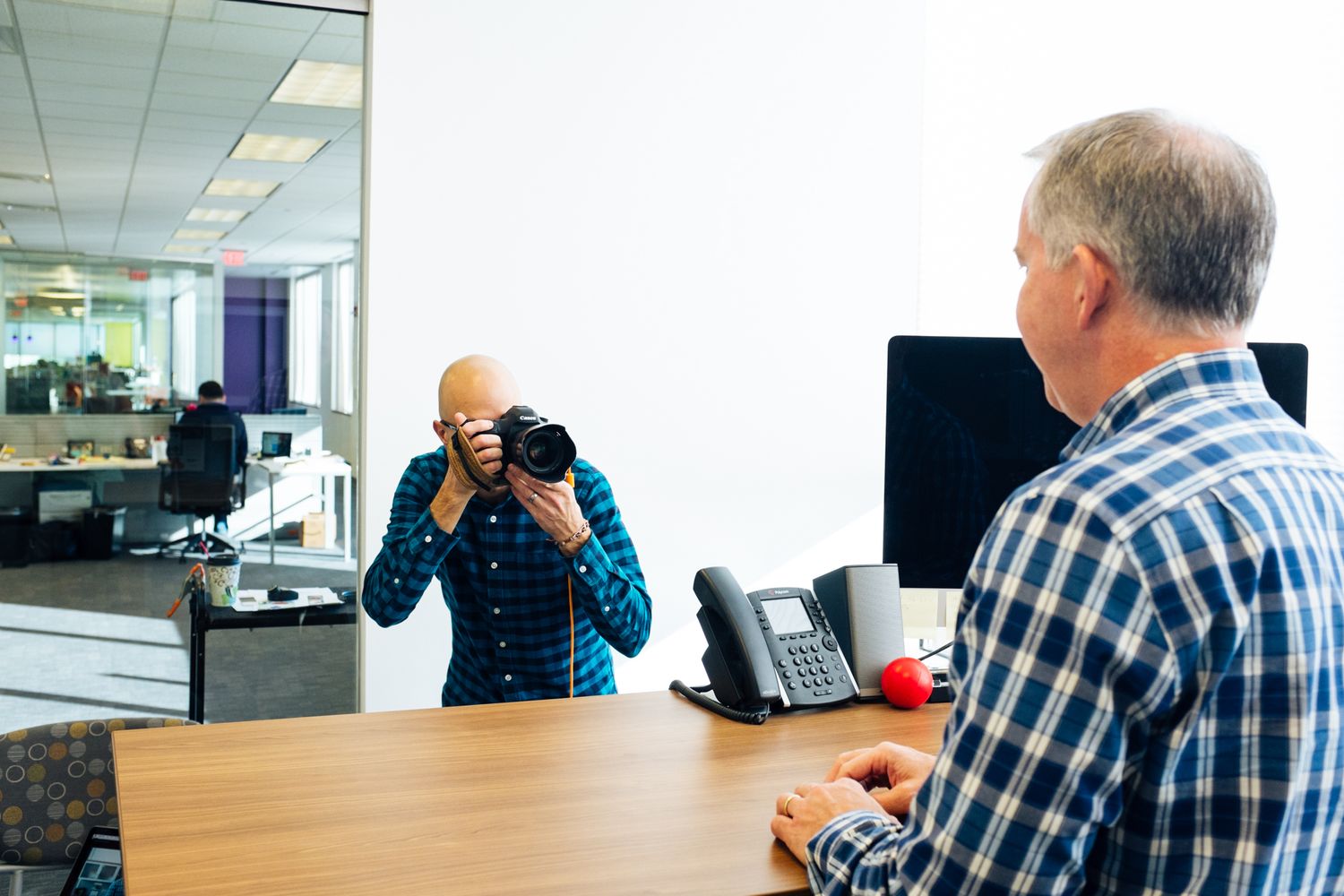 Photographer taking a portrait photo of a man sitting at an office desk with a DSLR camera.