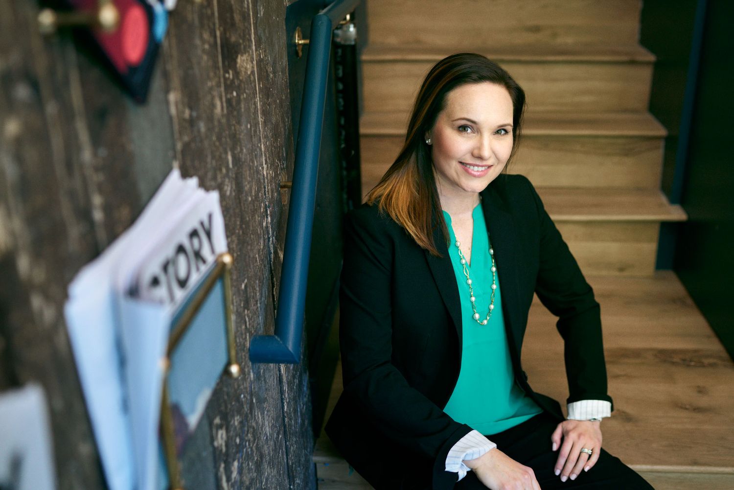 Smiling businesswoman in black blazer and teal top sitting casually on stairs in a creative office environment.