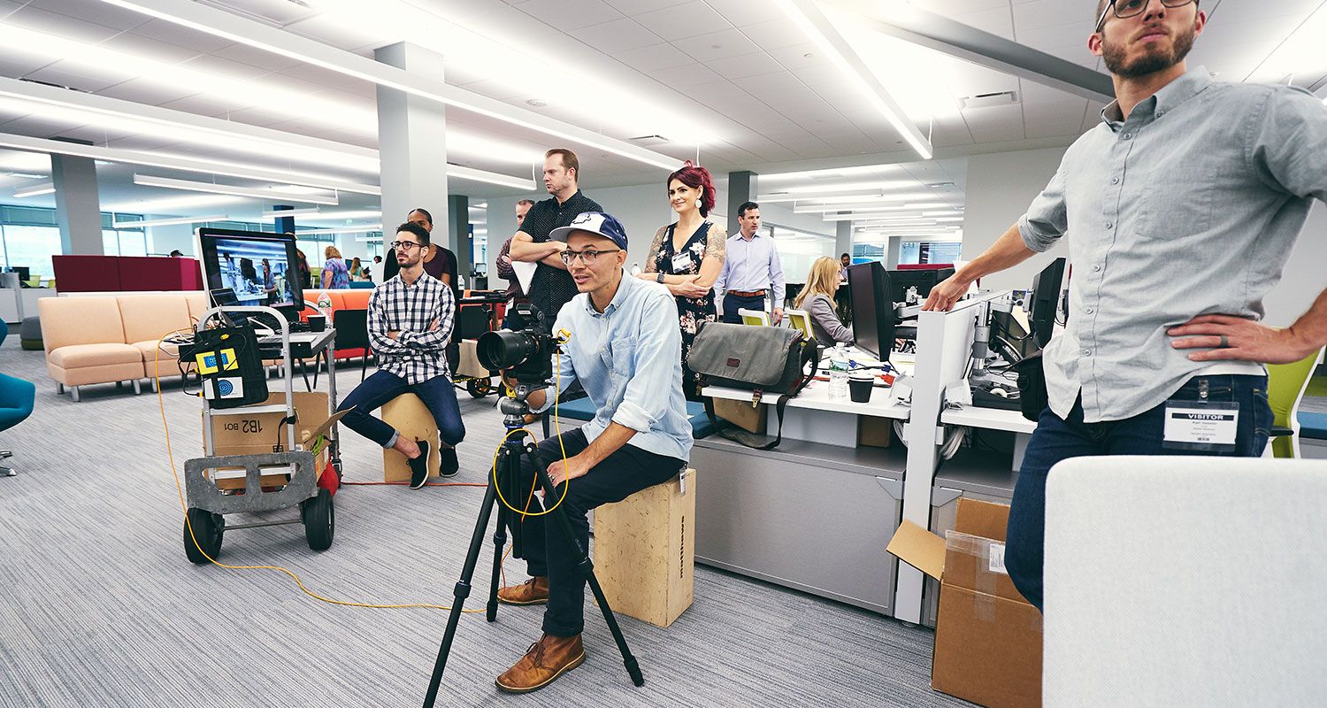 Camera crew filming in a modern open-plan office with multiple team members and equipment.