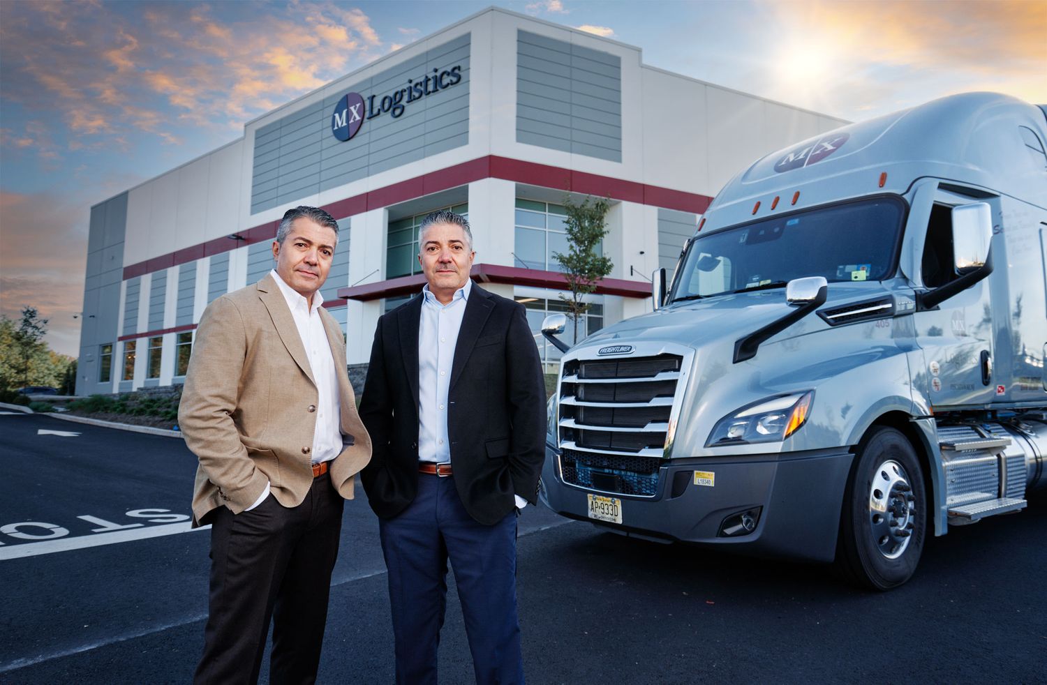 Two businessmen stand confidently in front of logistics facility and semi-truck at sunset.