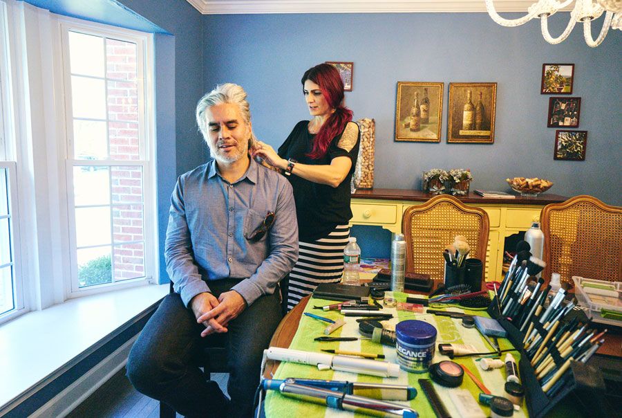 Red-haired makeup artist applying makeup to seated man near table with professional beauty tools.