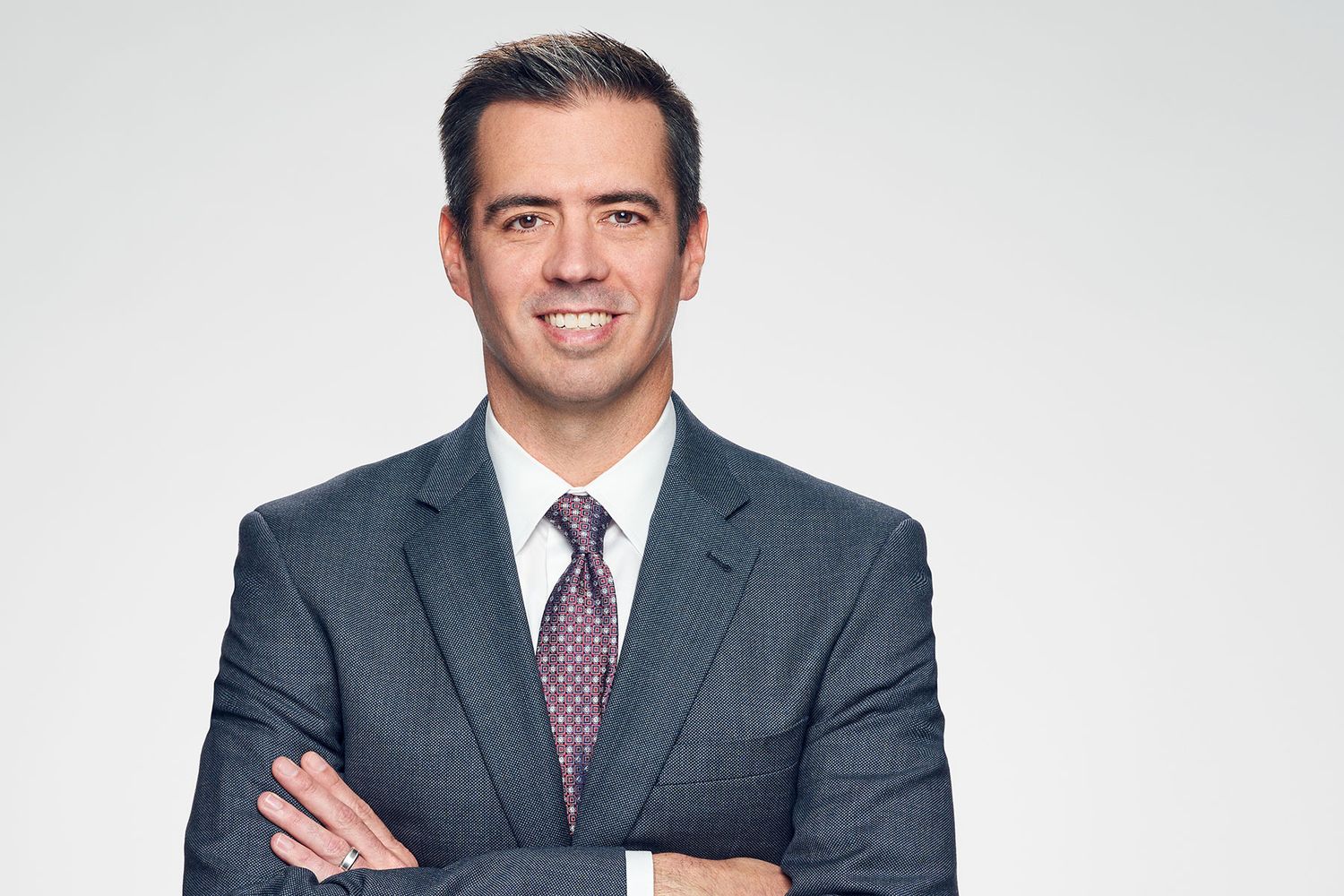Professional businessman in gray suit and tie smiling with arms crossed against white background.